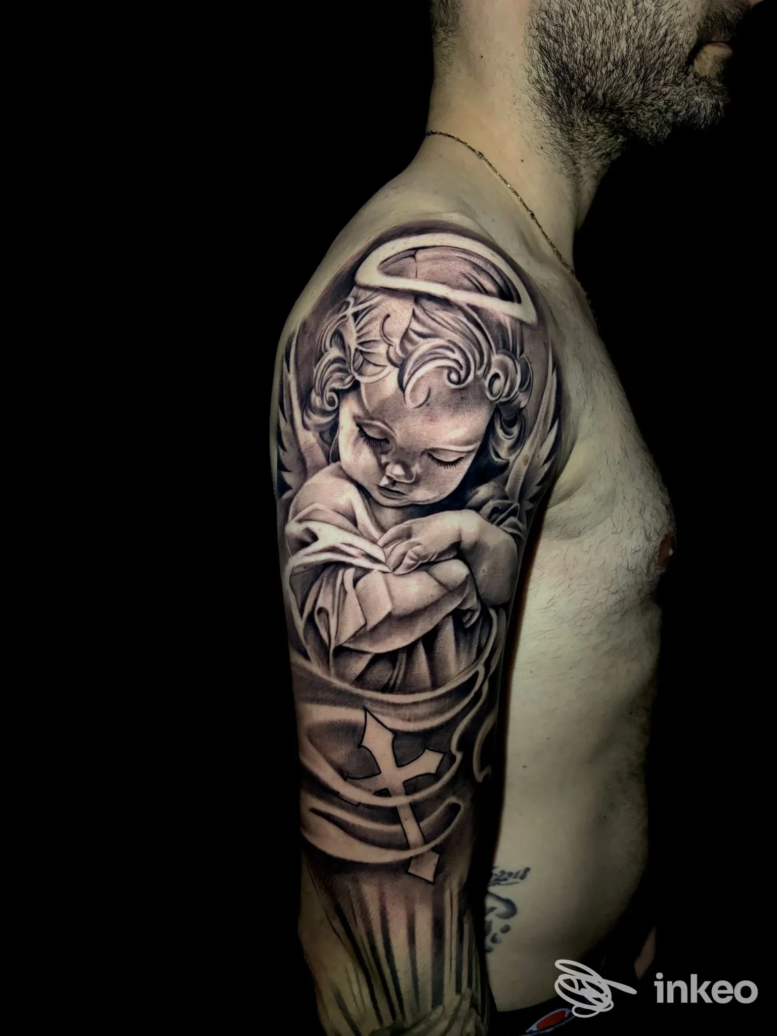 Tattoo realistico sul braccio sinistro di un uomo: angelo bambino in bianco e nero, contorni delicati e sfumature di grigio, sfondo scuro; realizzato dallo studio Doca Tattoo, Gioia Tauro.