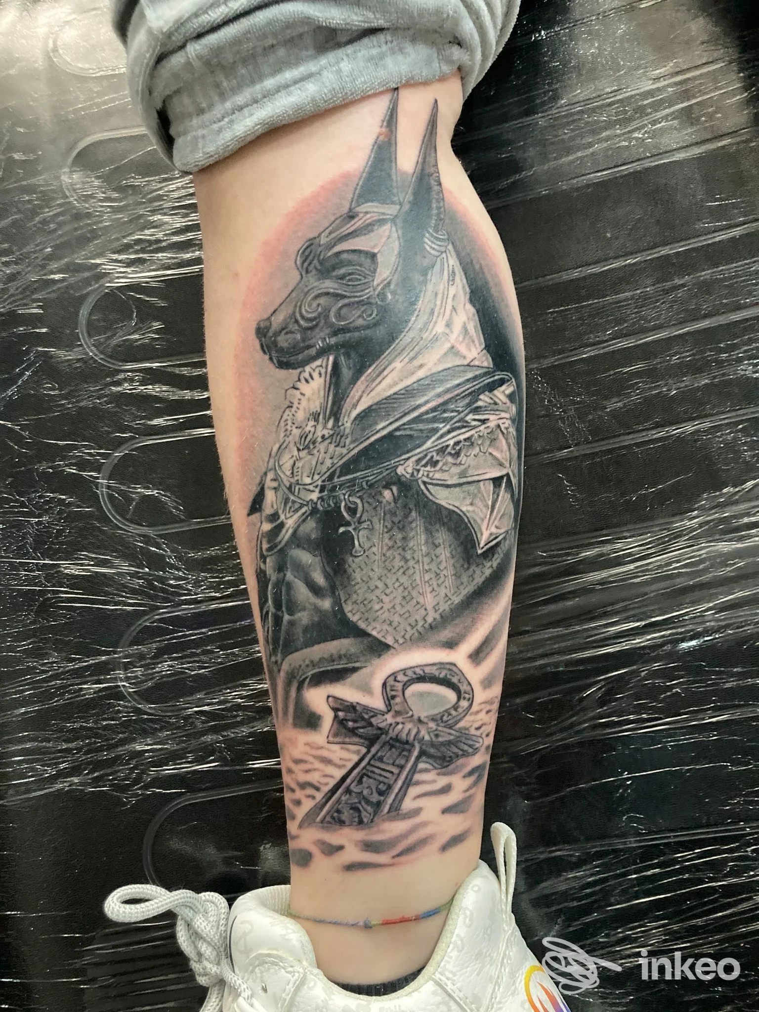 Tattoo visibile sullo svòrgio del braccio sinistro: un drago o creatura alata in bianco e nero, con dettagli realistici e sfondo sfumato; in basso si intuisce una croce egizia o un simbolo simile. l’immagine mostra una pelle reale tatuata senza elementi di ritocco digitale.