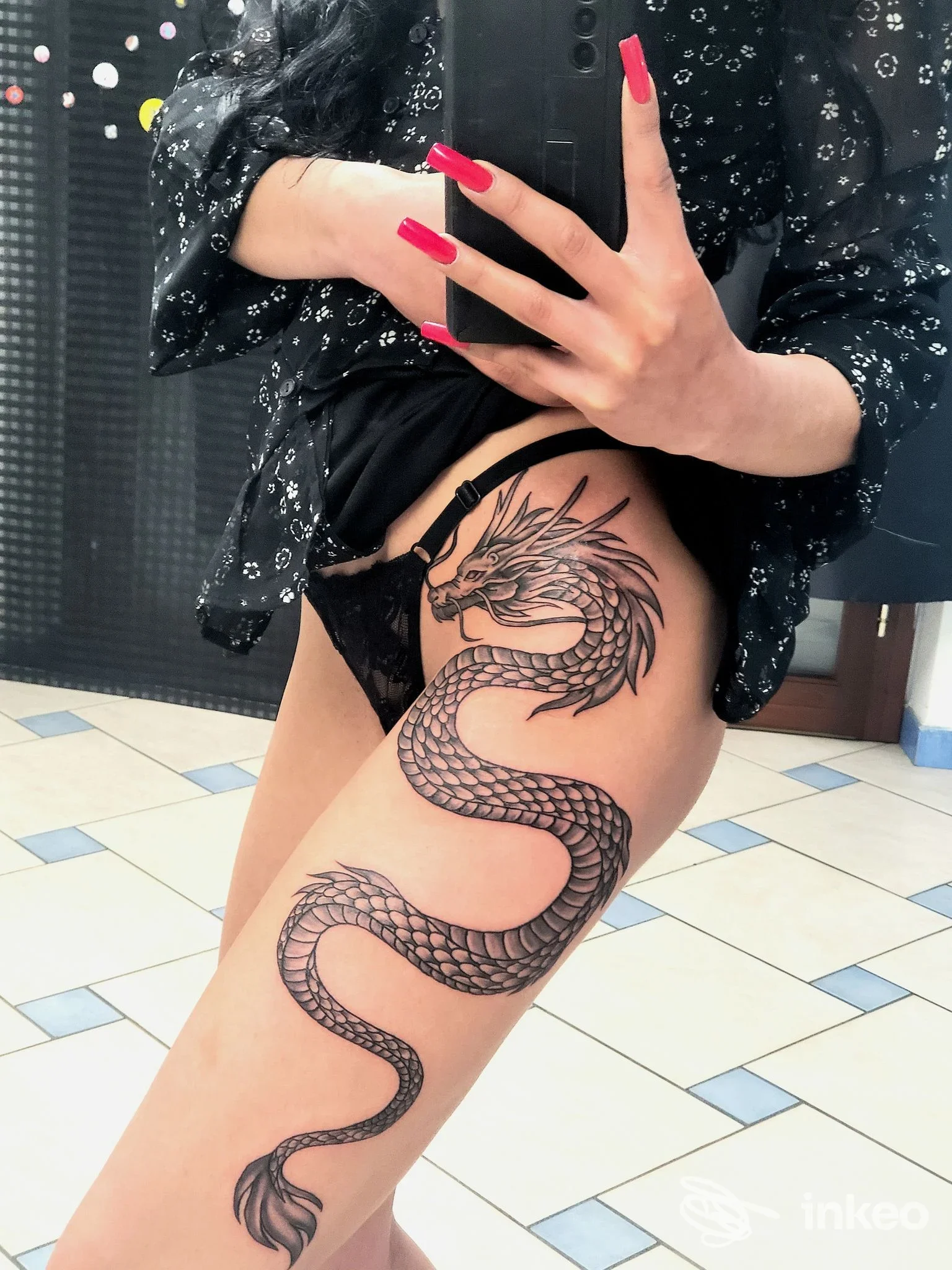 Tatuaggio corporeo in gamba raffigurante serpente/dragone in stile linework su coscia, visibile in primo piano con nastro in pizzo e dita lambite dal telefono, senza volto visibile.