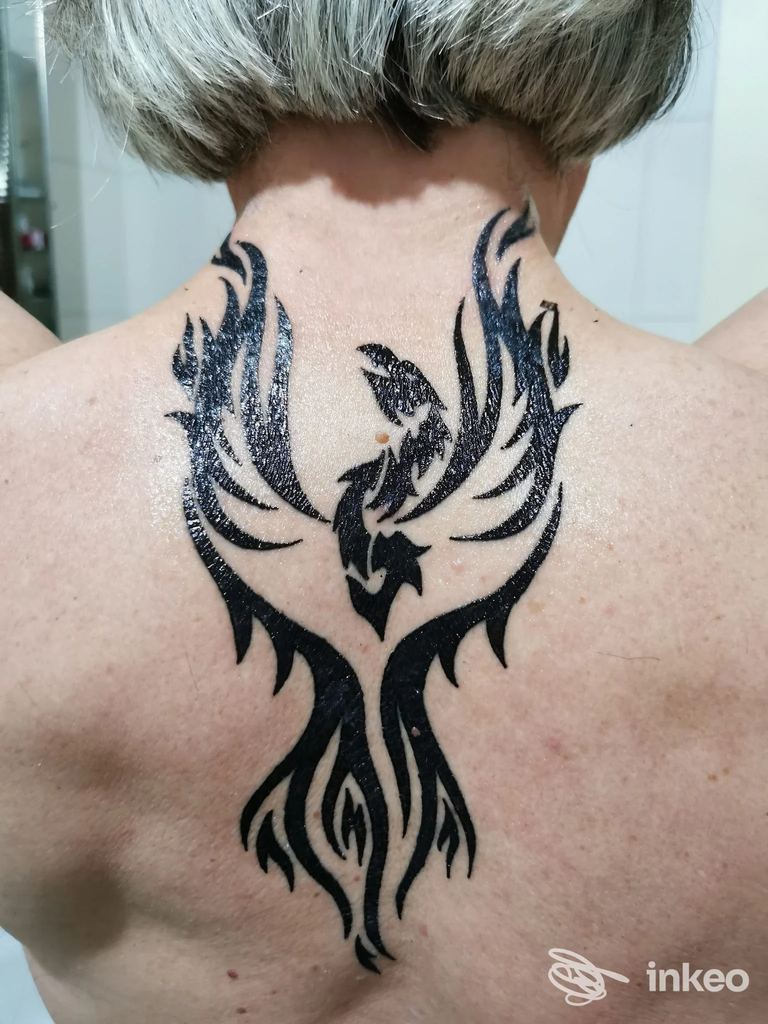 Tatuaggio a sfondo nero a forma di fenice stilizzato sulla parte alta della schiena, visibile completamente e ben definito; corpo senza volto visibile, studio Art Tattoo Damiano, Corigliano Scalo.