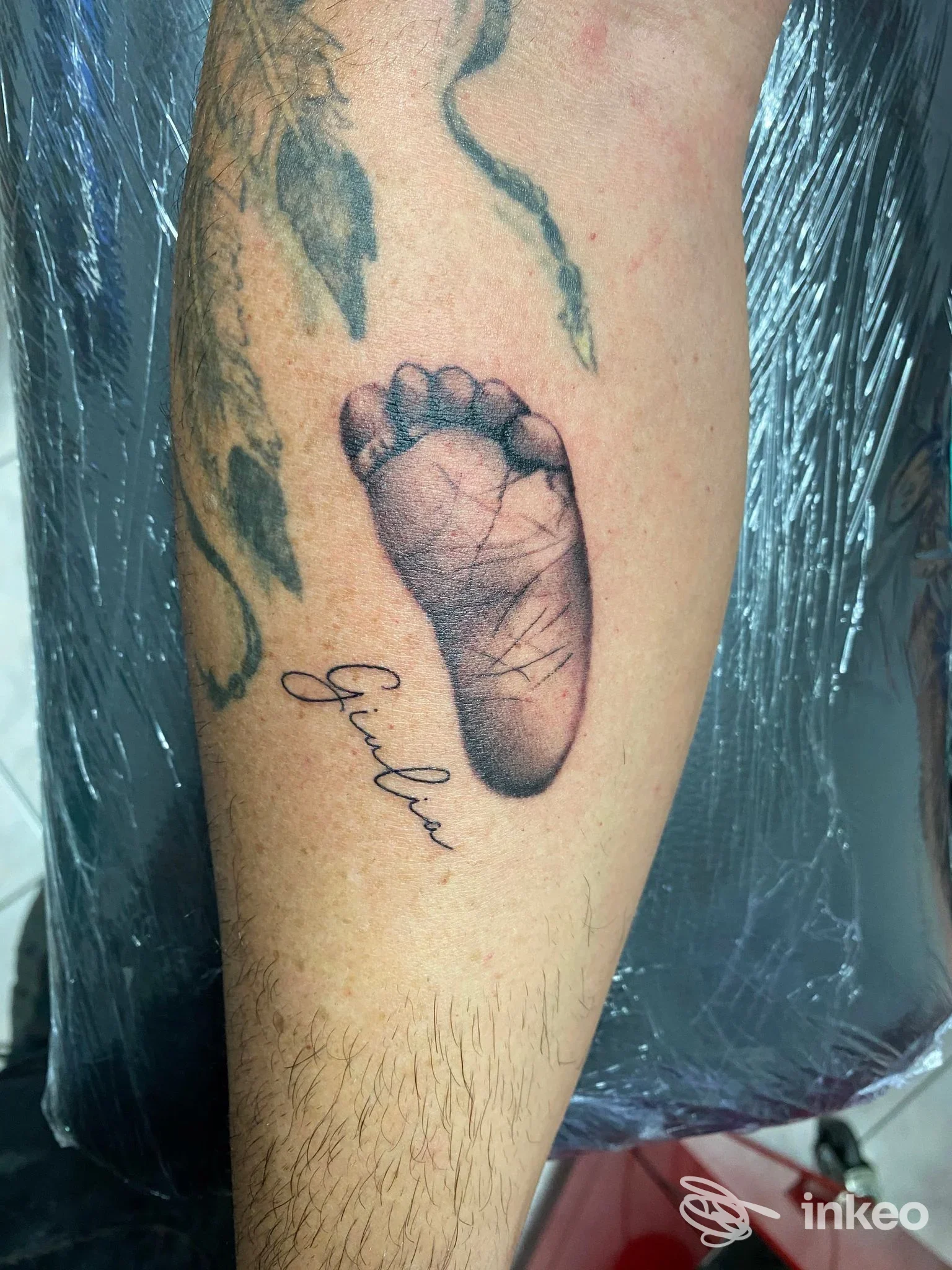 Applet di gamba con impronta di piede realistica e nome in corsivo scritto accanto, tatuaggio chiaramente visibile sulla pelle.
