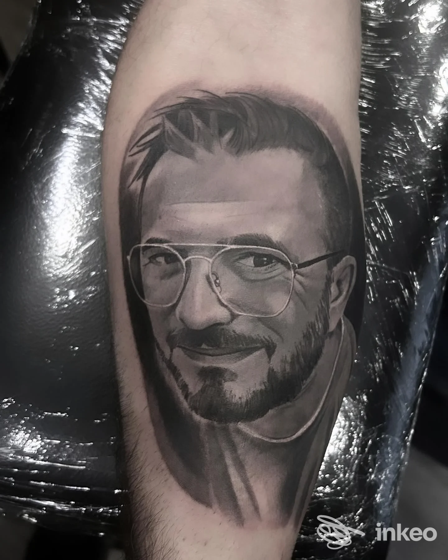 Tattoo realistico su avambraccio raffigurante una persona con occhiali, eseguito su pelle reale, colori nero/grigi.