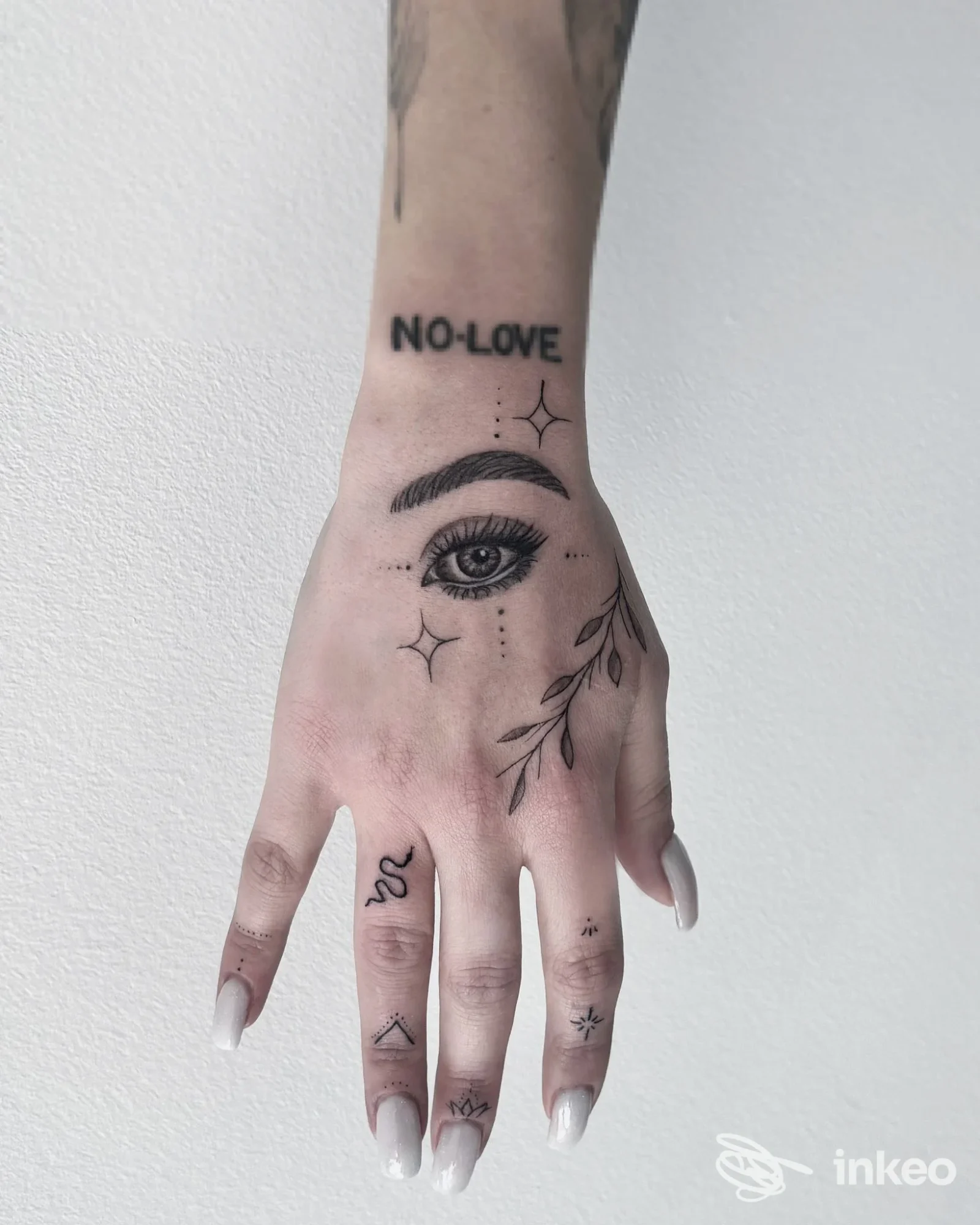 Schermata di mano con tatuaggio: occhio centrale, decorazioni stellate e rami, scritta NO-LOVE sul polso, senza presenza visibile del volto.