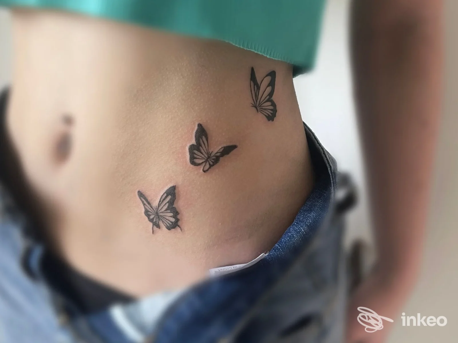 Dettaglio di un tattoo di tre farfalle nere sulla pelle della zona lombare/addominale di una persona, con jeans abbassati leggermente e una fascia verde sopra.