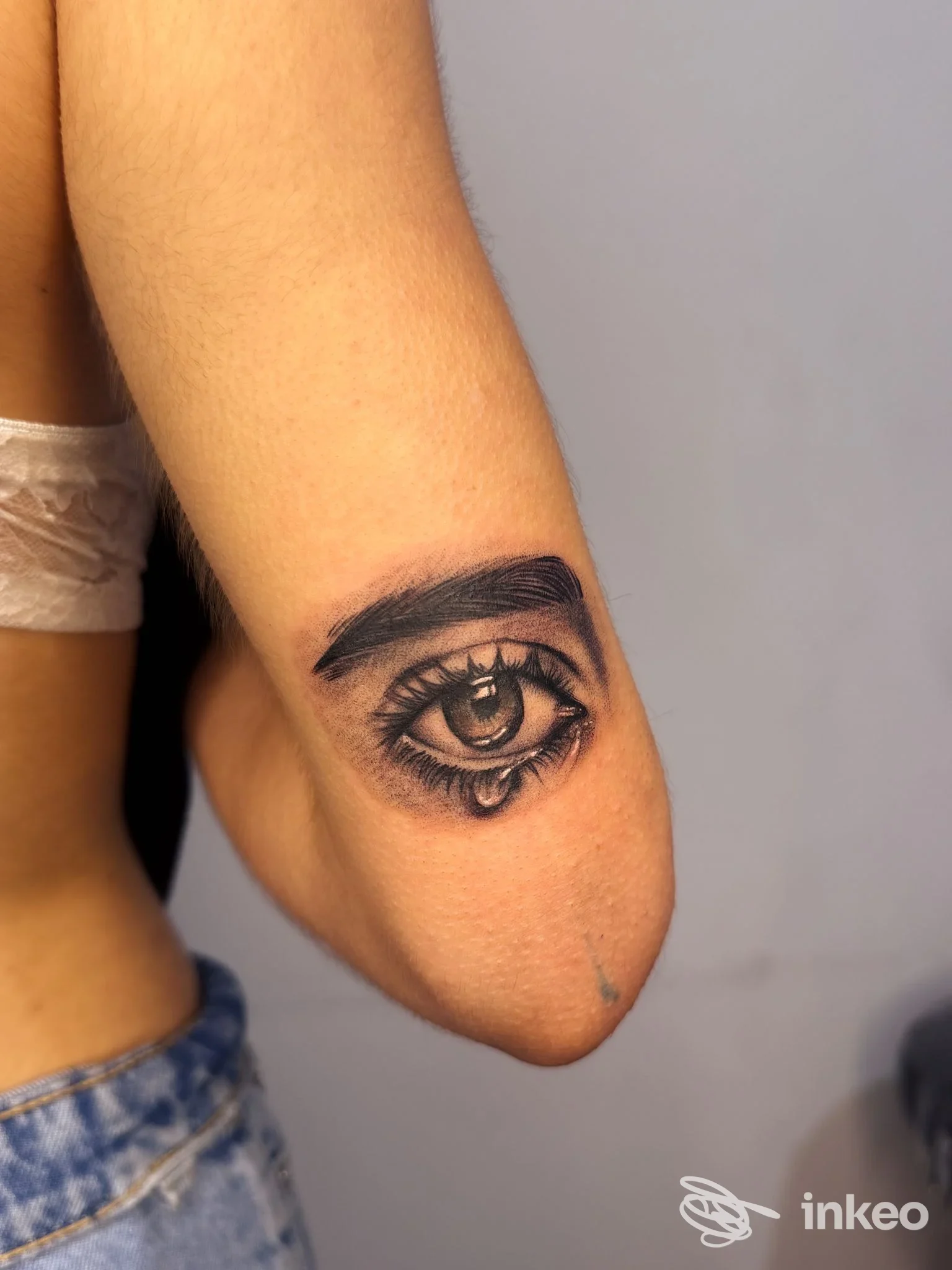 Tattoo realistico di un occhio posto sull’avambraccio, dettagli di ciglia e lacrima, in tonalità scure su pelle chiara, con sfondo neutro.