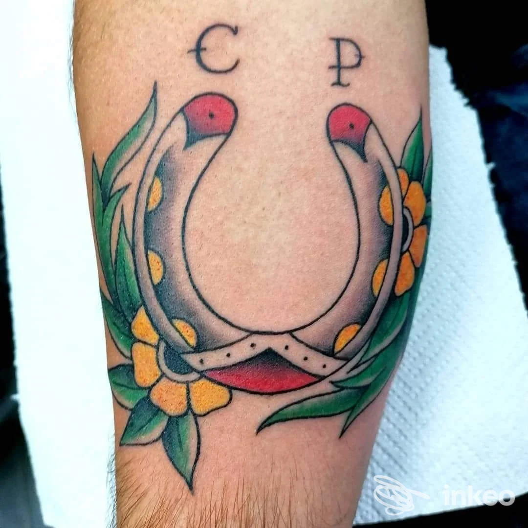 Tatuaggio visibile sull'avambraccio raffigurante una forma a uncino decorata con fiori gialli e foglie verdi, con dettagli neri e rossi.