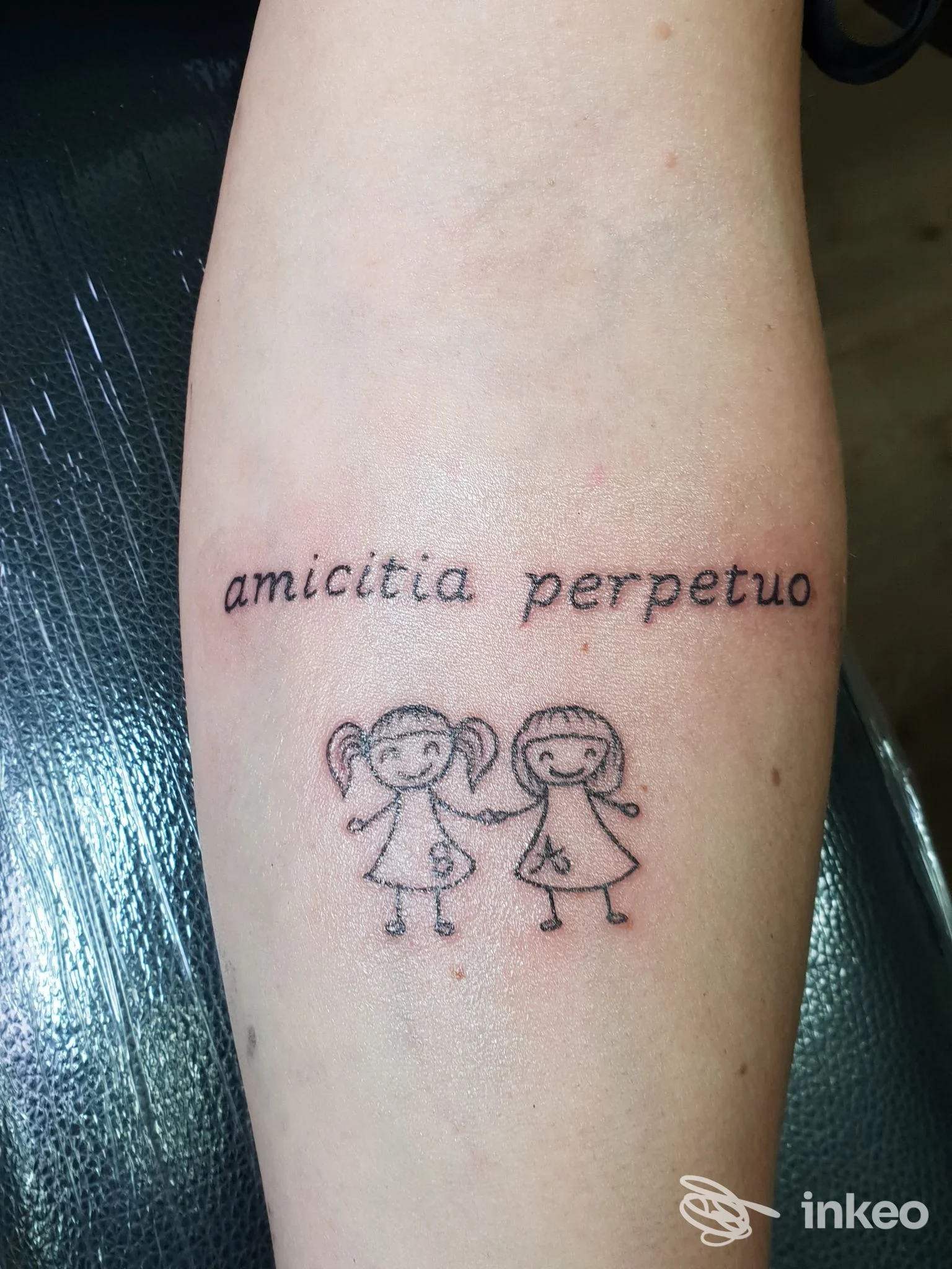 Tattoo sull'avambraccio raffigurante due bambine stilizzate che si tengono per mano, accompagnato dalla scritta latina 'amicitia perpetuo'.