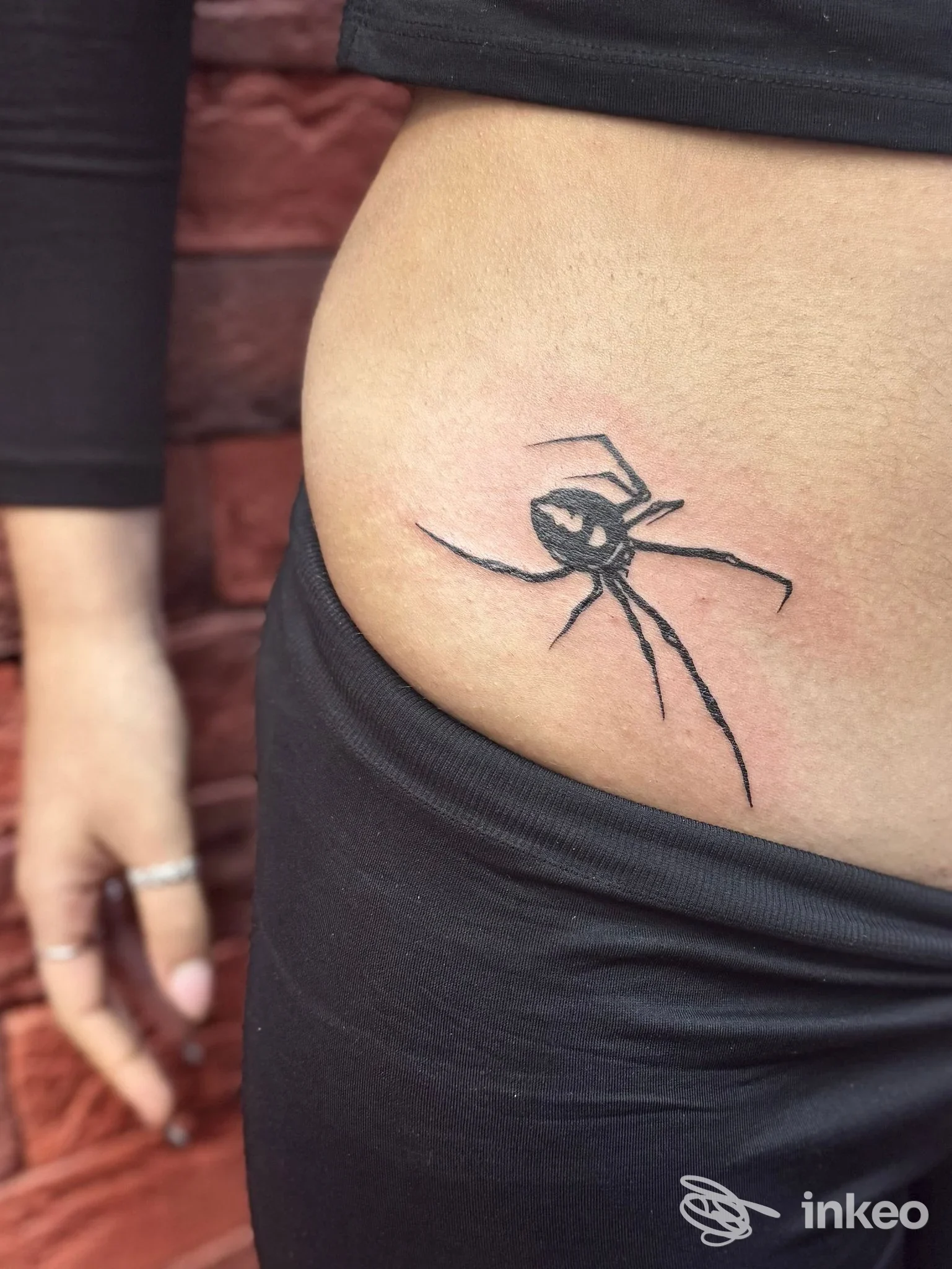 Tattoo realistico di ragno stilizzato in nero su pelle laterale dell'addome/inguine, vicino alla cintura dei pantaloni, eseguito su una persona.