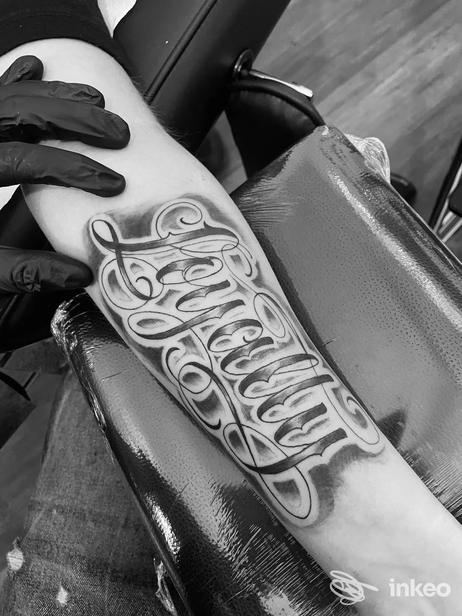 Tatuaggio in stile script calligrafico nero sull'avambraccio, ritratto da una mano guantata. nessun volto visibile.