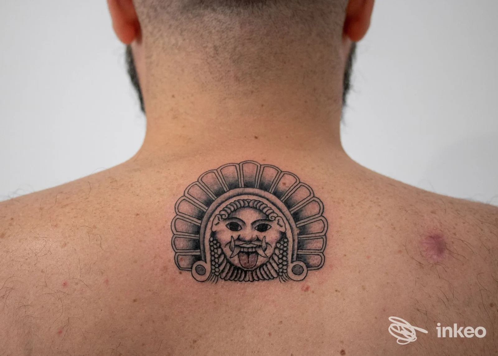 Tatuaggio sul dorso della nuca raffigurante un volto stilizzato con decorazione a elmo, eseguito a linework nero su pelle chiara.