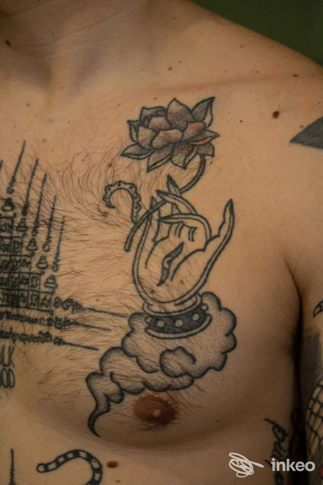 Torace di uomo con tatuaggio visibile: una mano che sostiene un bouquet di fiori, con ornamenti e nuvole sullo sfondo.