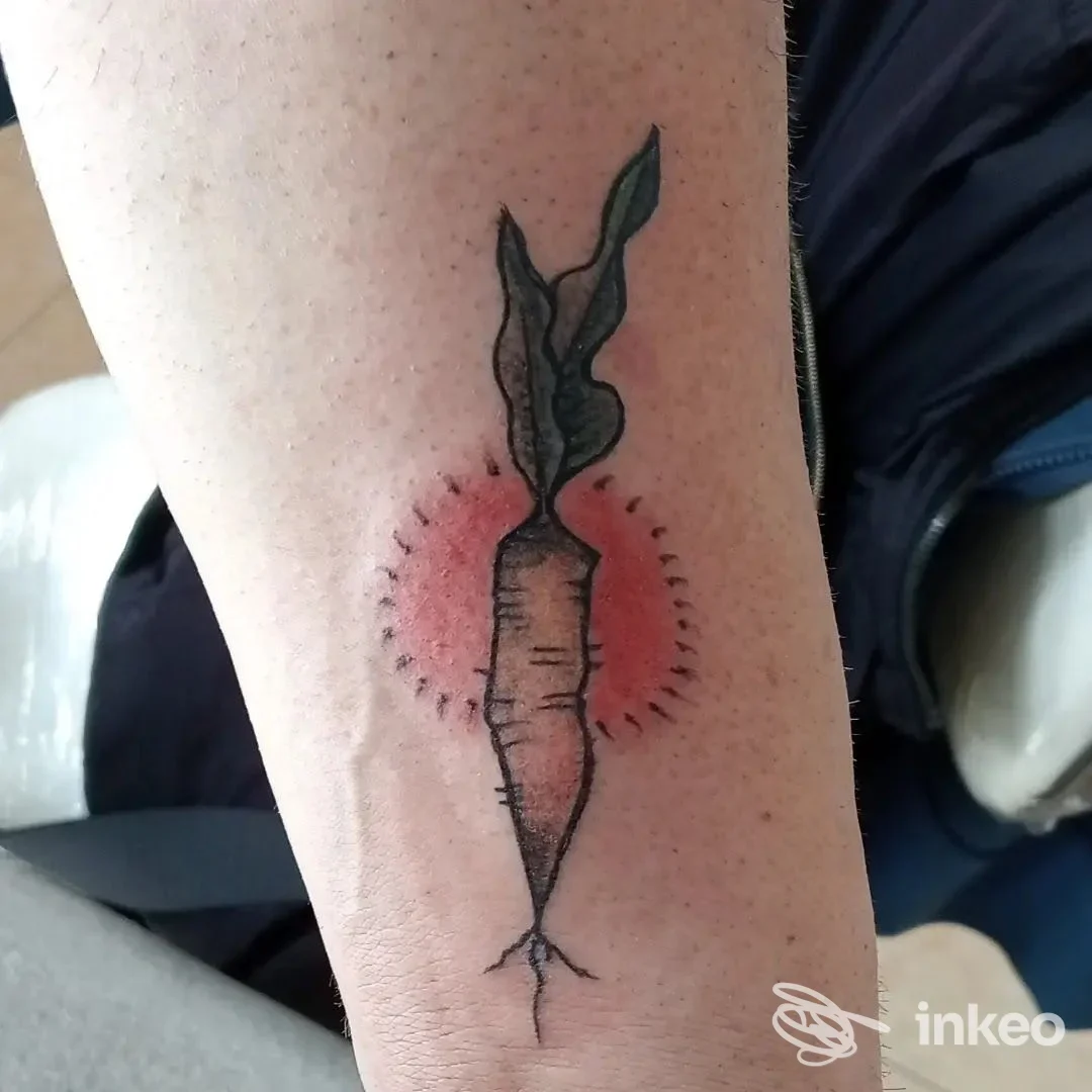 Tattoo raffigurante una carota stilizzata con foglie, eseguita sulla pelle della gamba, con sfondo rosato e contorno scuro.