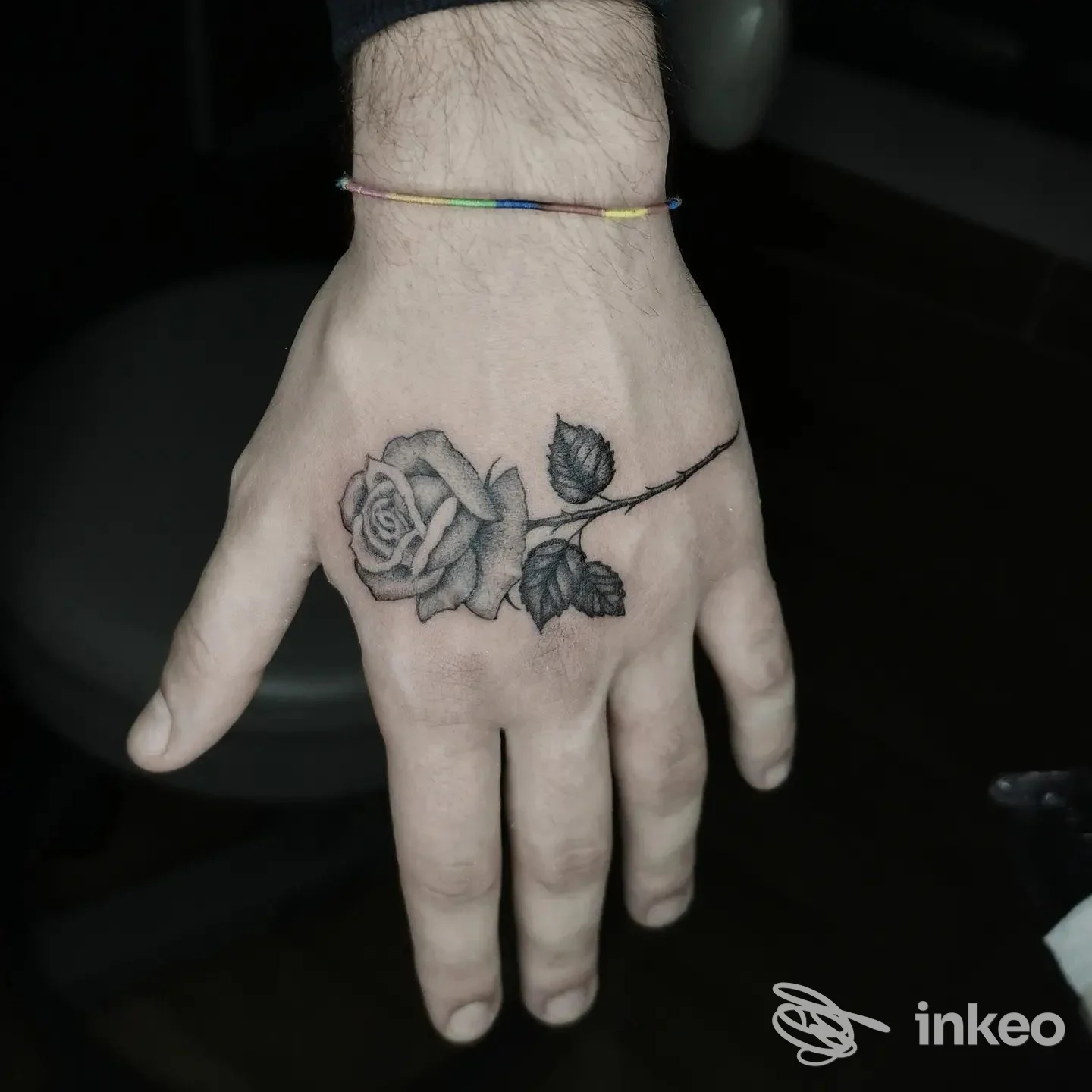 Tatuaggio realistico di rosa con stelo sul dorso della mano, tonalità nero/grigio, accompagnato da un piccolo braccialetto multicolore al polso.