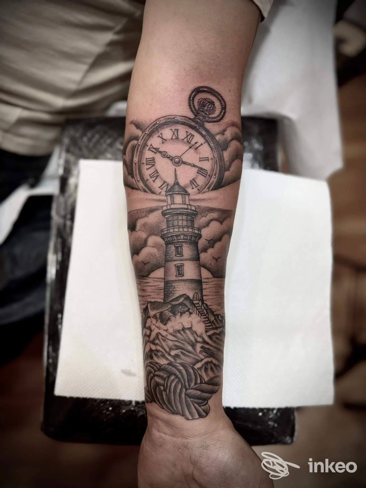 Tattoo sull'avambraccio con un faro circondato da nuvole e un orologio tascabile: chiaro disegno in bianco e nero su pelle, nessun volto visibile.