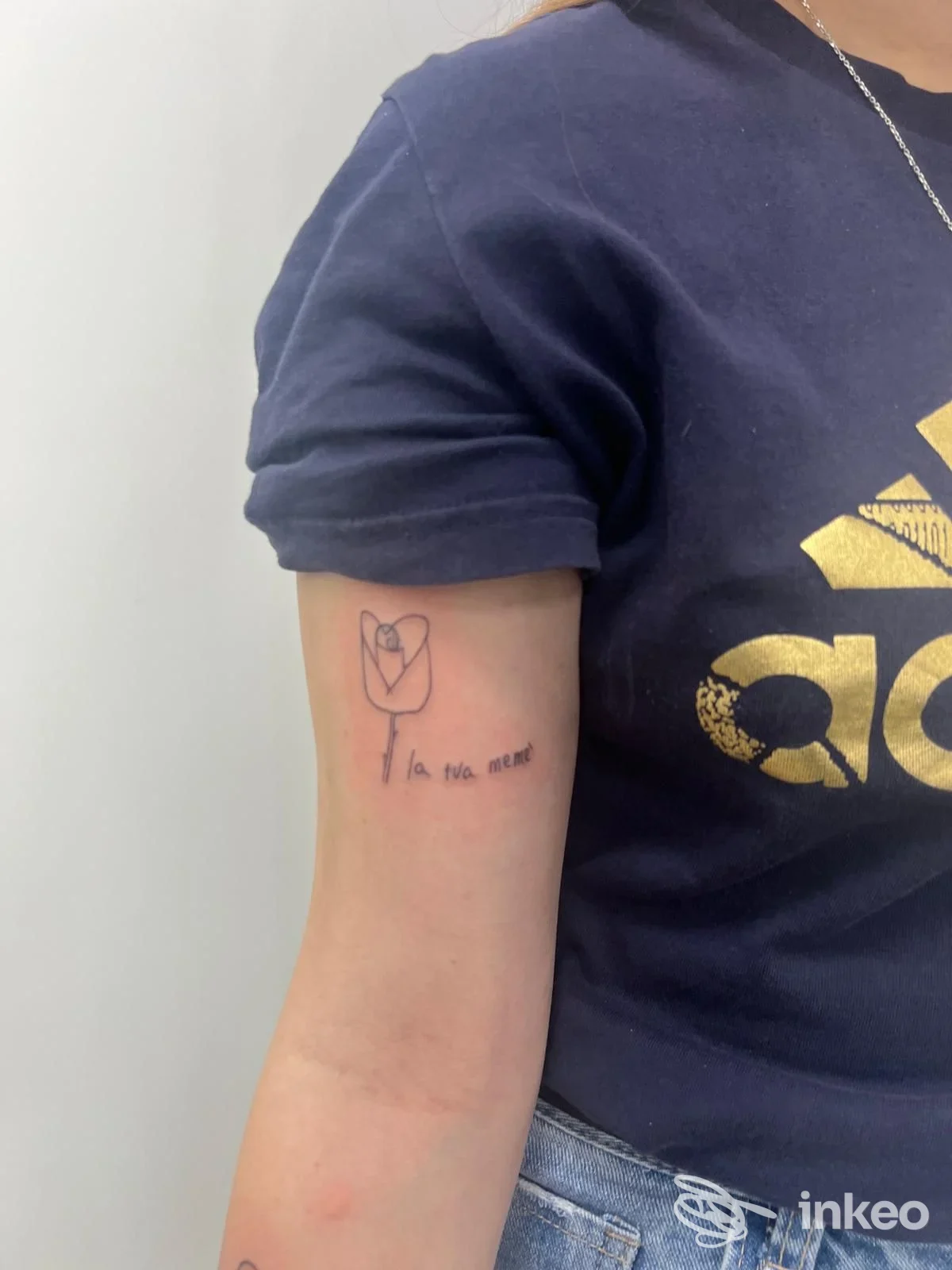 Tattoo visibile sull’avambraccio sinistro: disegno semplice di fiore/cuore con testo accanto, tatuaggio su pelle nuda, senza volto visibile nella foto.