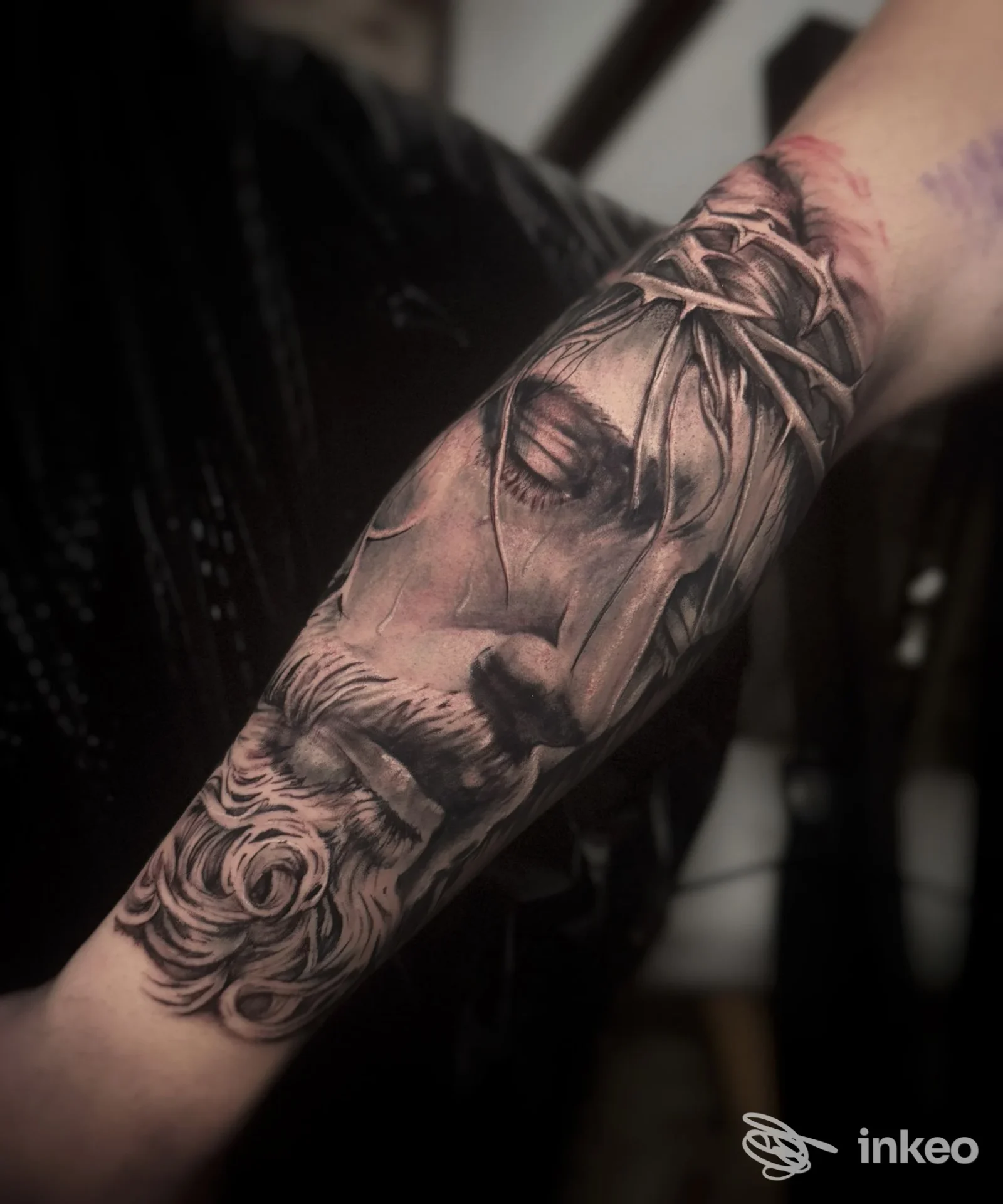 Braccio di una persona con tatuaggio realistico in bianco e nero di un volto e corona di spine, sfumato e dettagliato.