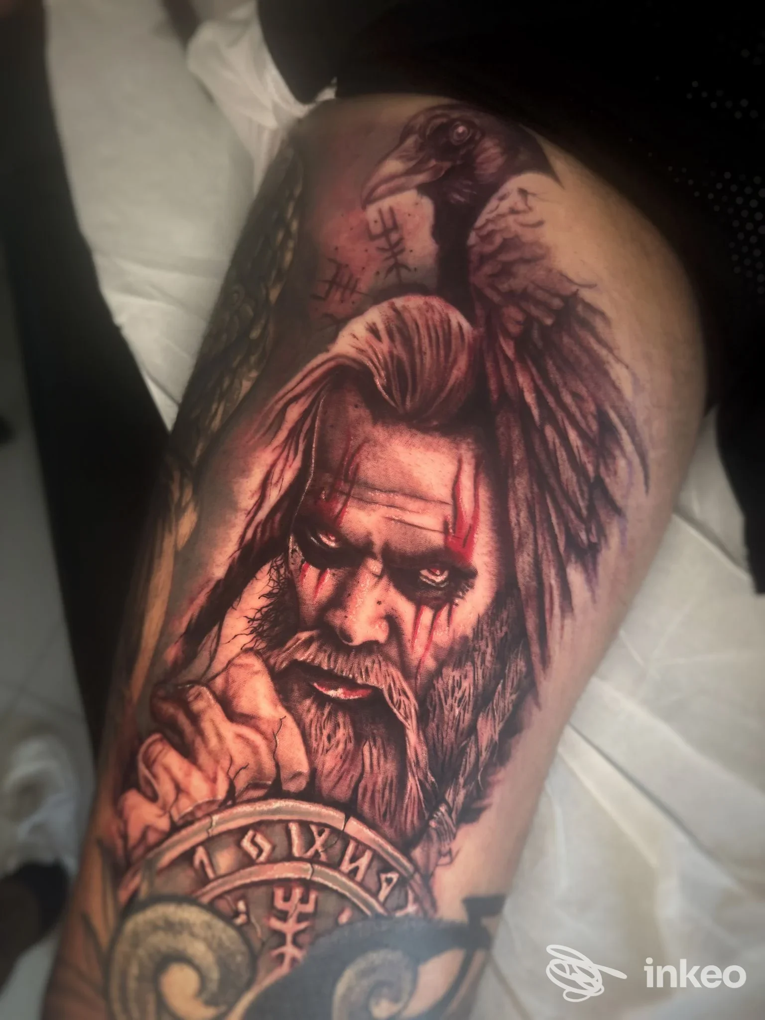 Tattoo realistico su braccio raffigurante una figura barbuta di fronte, con dettagli drammatici e simboli circolari, in tonalità scure su pelle.