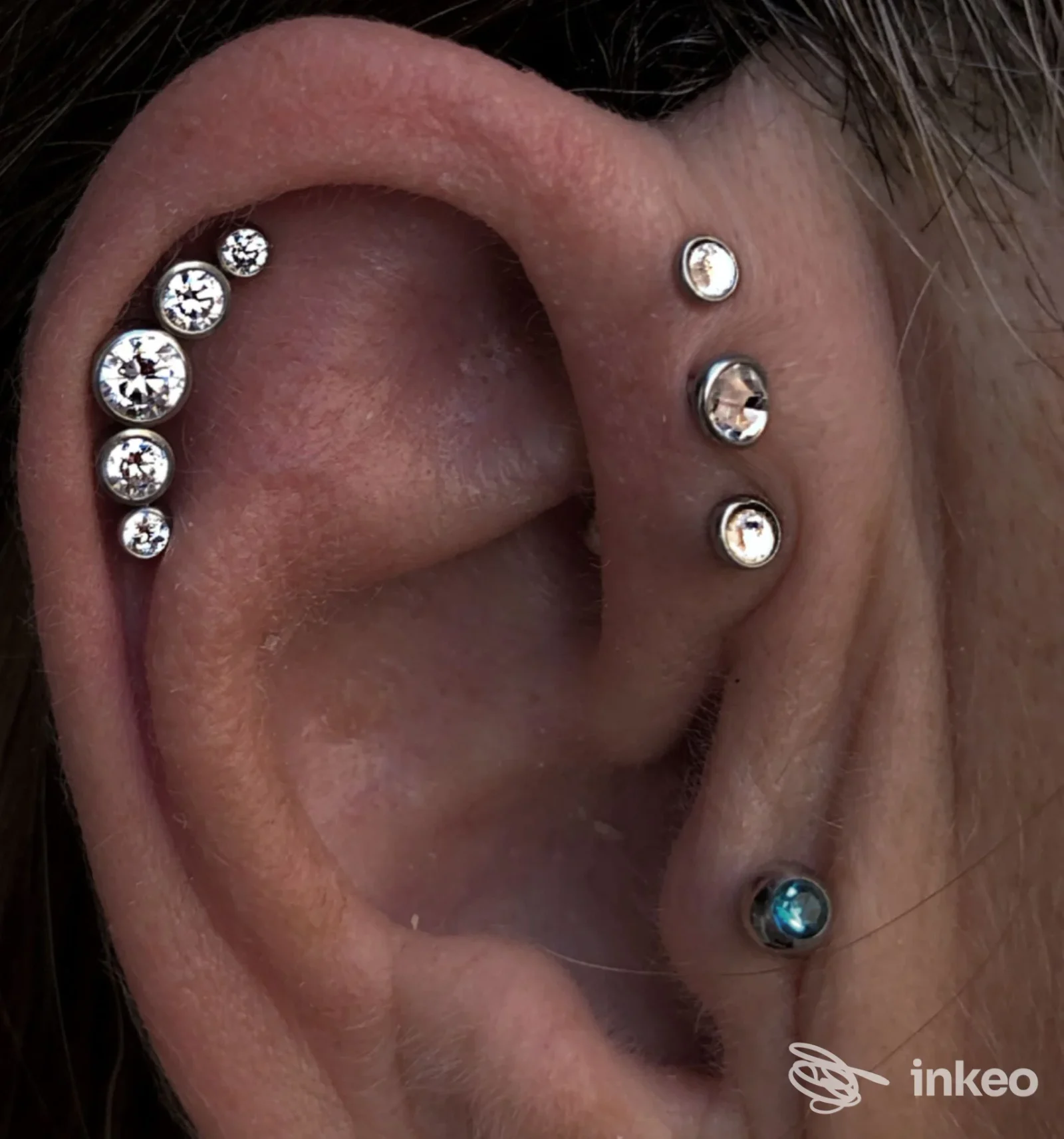 Primo piano di un orecchio con piercing multipli: una fila di gemme argentate lungo la conca e piercing aggiuntivi lungo la piega esterna, realizzati sulla pelle.