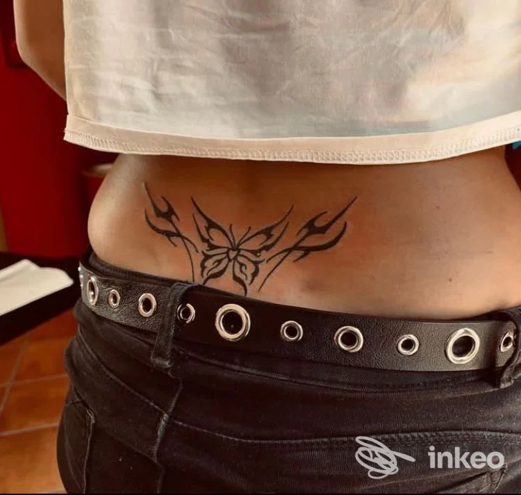 Tattoo nero sul fianco/basso dorso, design stilizzato a farfalla o fiore, visibile su pelle nuda, cintura in vita con borchie.