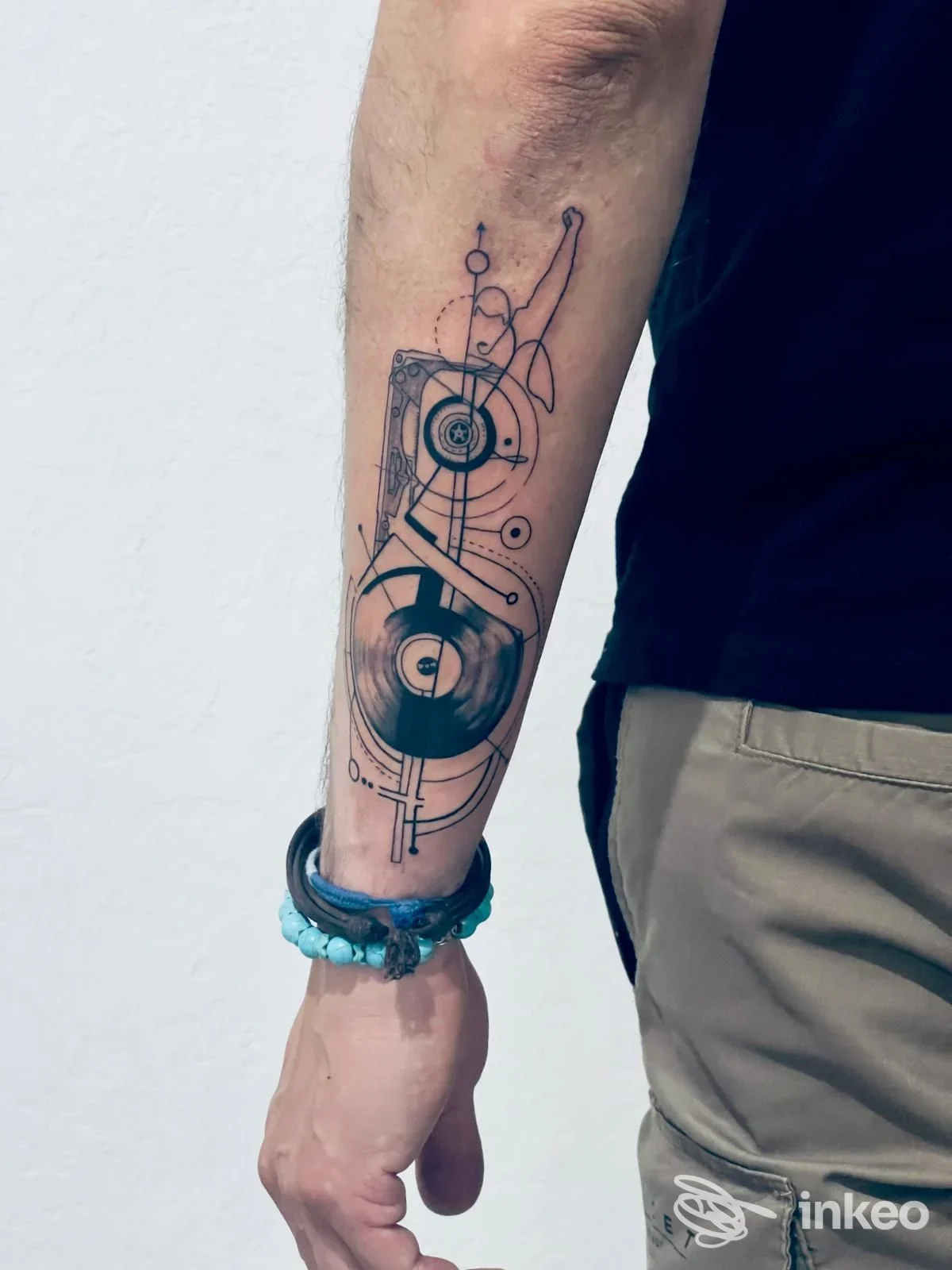 Avambraccio di una persona con tatuaggio geometrico e circolari su linee nere, stile tecnico, braccialetto azzurro al polso, sfondo bianco.
