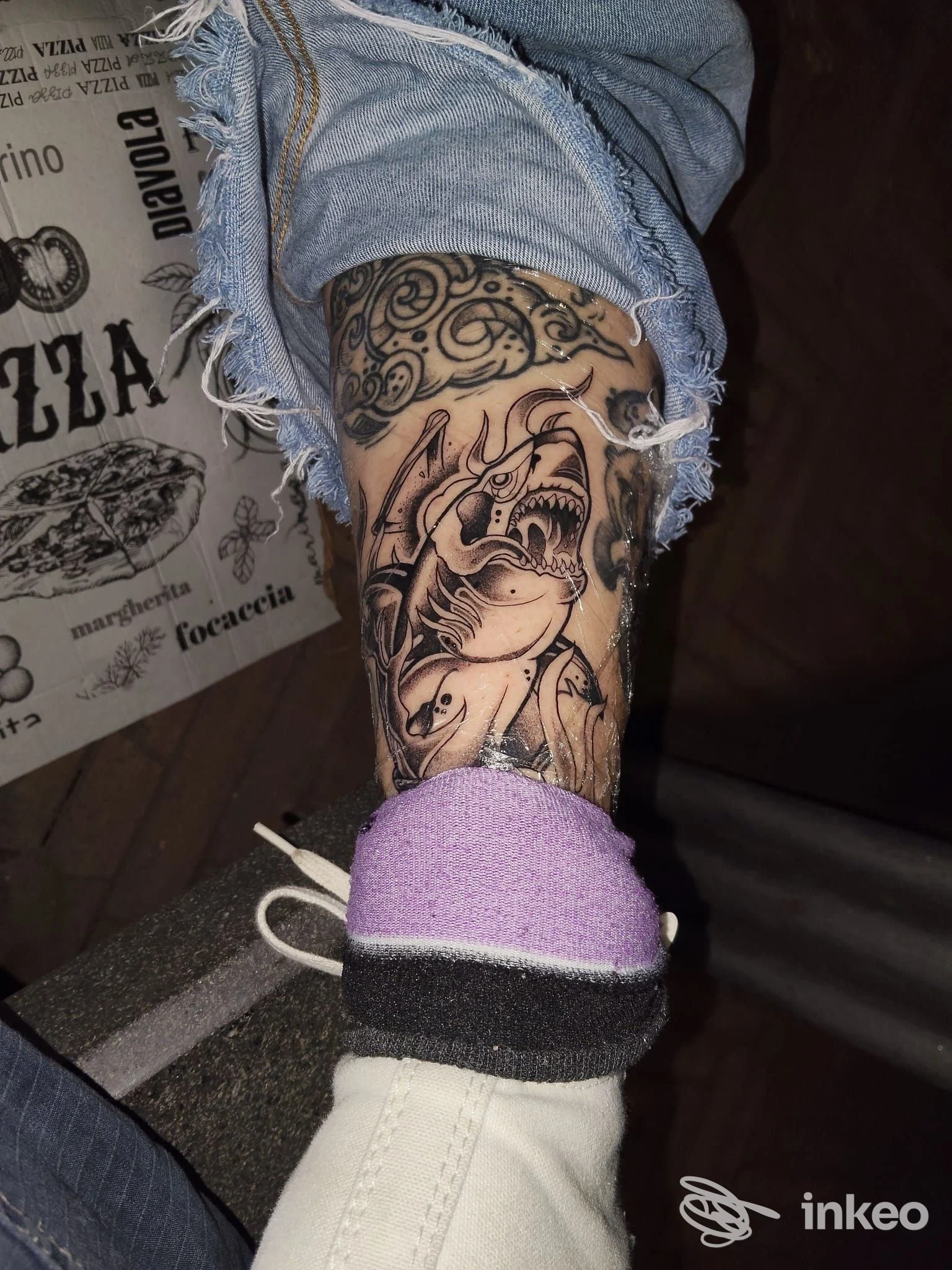 Tattoo completo su polpaccio di una persona, disegno ornamentale in stile linework con sfumature e contorni scuri, indossando pantaloni strappati e una sneaker viola.