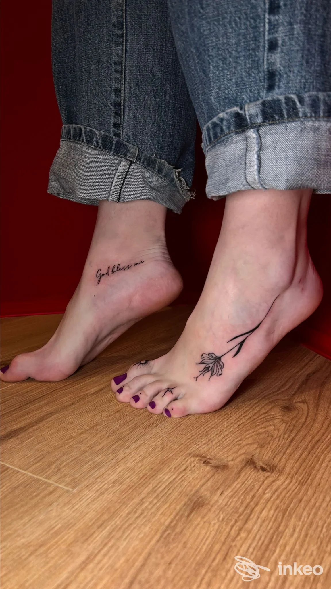 Due piedi nudi con tatuaggi visibili: una scritta sul tallone sinistro e un fiore stilizzato sul dorso del piede destro, indossano jeans strappati, sfondo rosso e pavimento in legno.