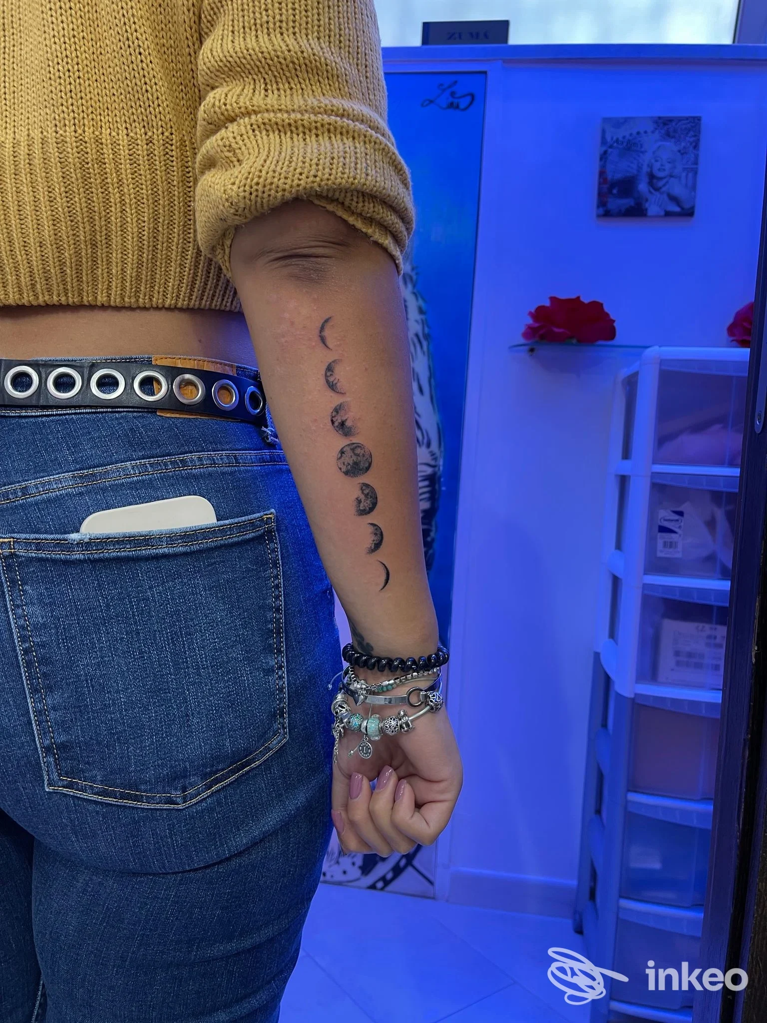 Braccio di una persona con una sequenza di fasi lunari tatuate lungo l'interno dell'avambraccio, indossa maglia beige e bracciali, sfondo di studio tattoo.