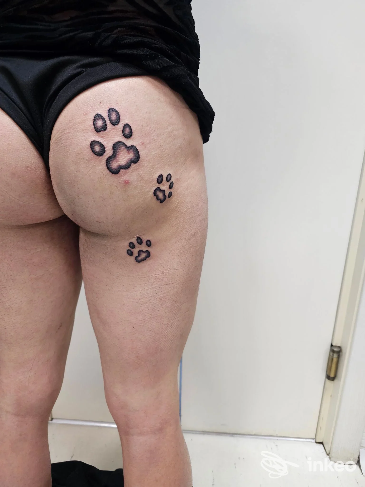 Tattoo su gluteo e parte alta della coscia: impronte di zampe nere con contorno grigio scuro. nessun volto visibile, sfondo chiaro.
