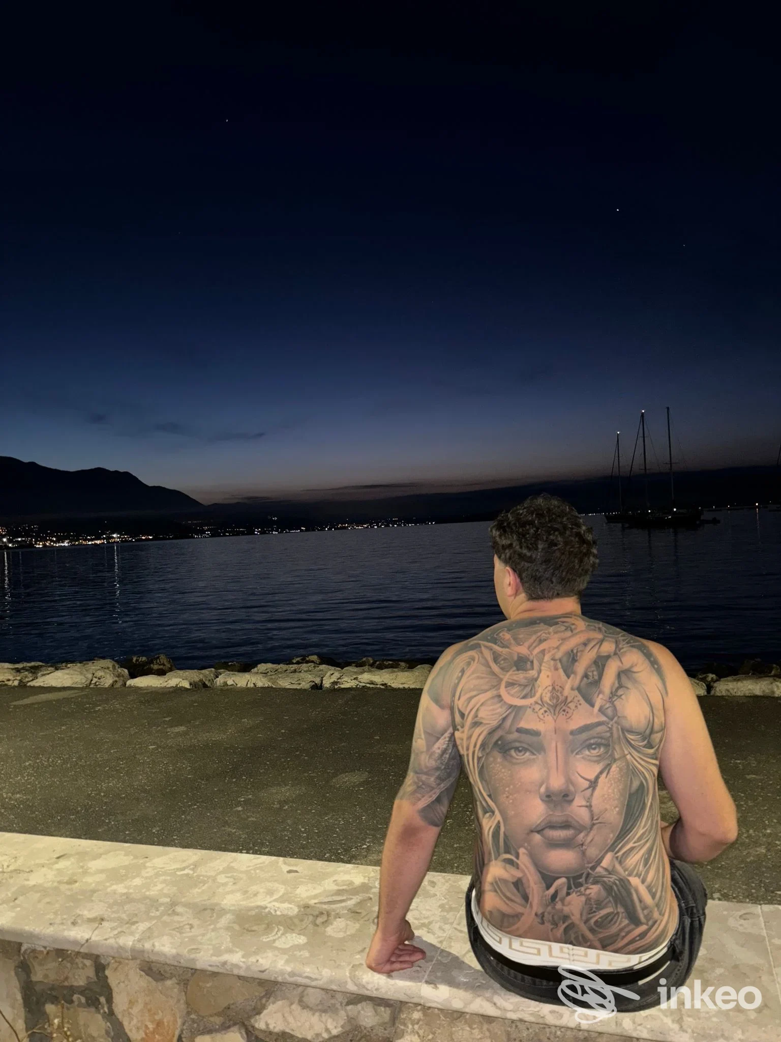 Una persona vista di schiena, seduta su una cornice al mare al crepuscolo: sul dorso è visibile un grande tatuaggio che copre quasi tutta la schiena.