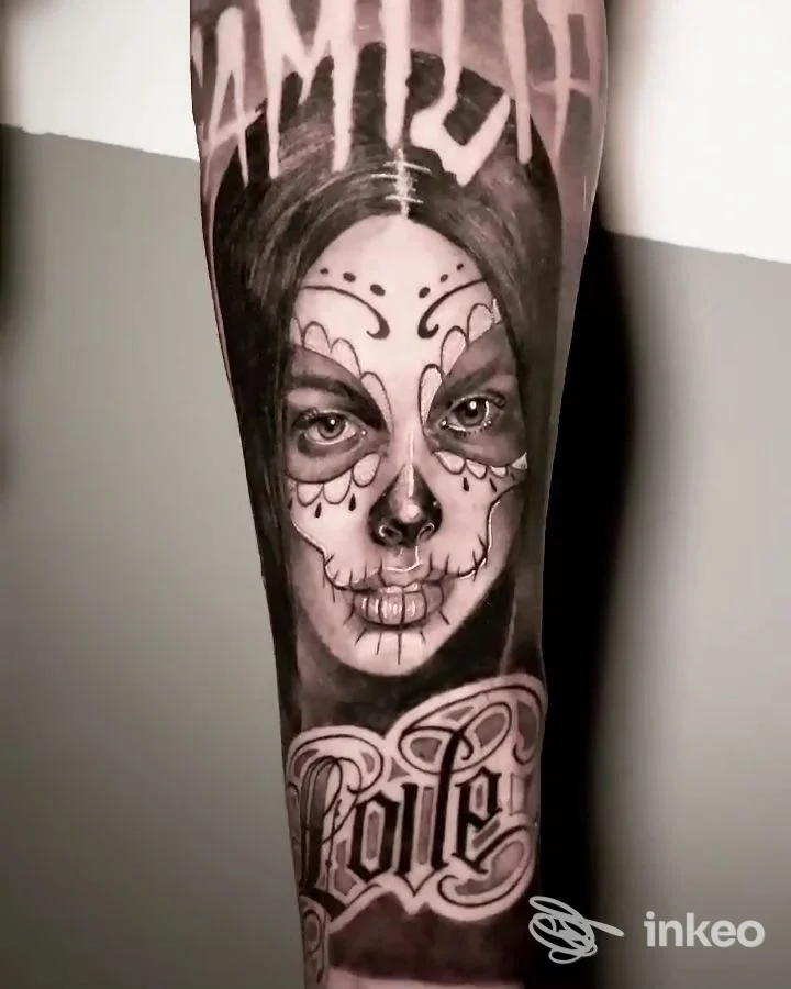 Braccio visibile con tatuaggio in stile Sugar Skull su volto femminile, dettagli scuri e scritte decorative, tonalità nero- grigio.
