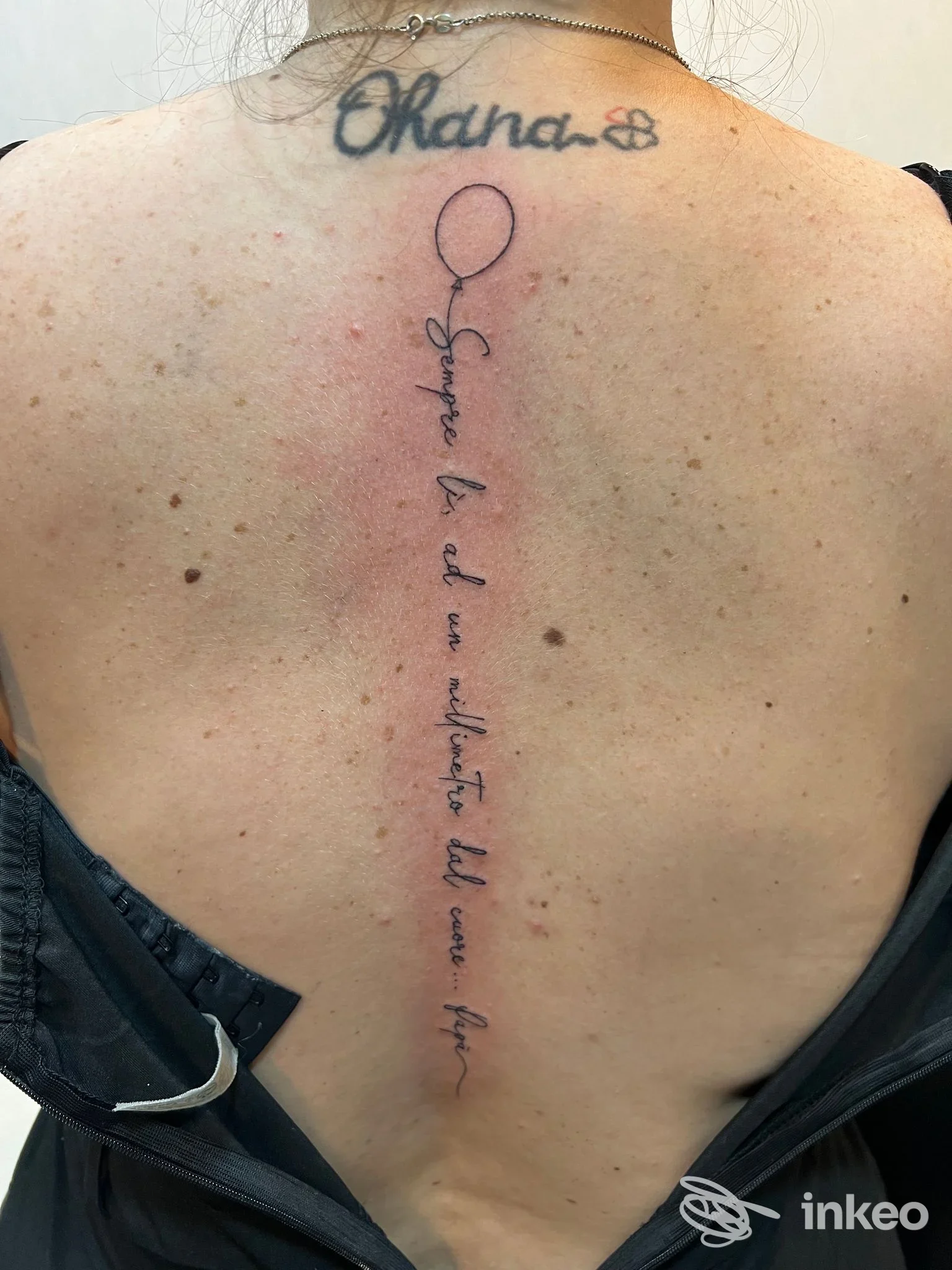 Tattoo lungo sulla schiena, testo in corsivo che corre verticalmente lungo la colonna vertebrale; in alto è presente un anello/loop che sembra un palloncino.