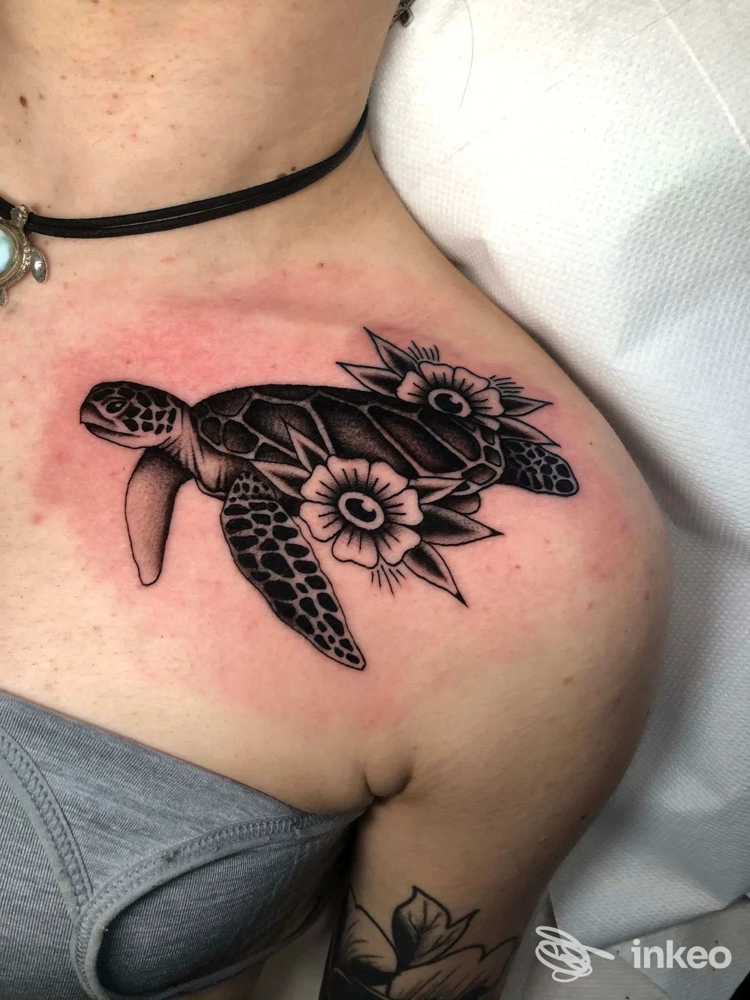Tartaruga tatuata inchiostrata in nero con fiori, applicata sulla spalla sinistra di una persona, con pelle leggermente arrossata attorno all'area. Nella foto si intravede una collana nera.