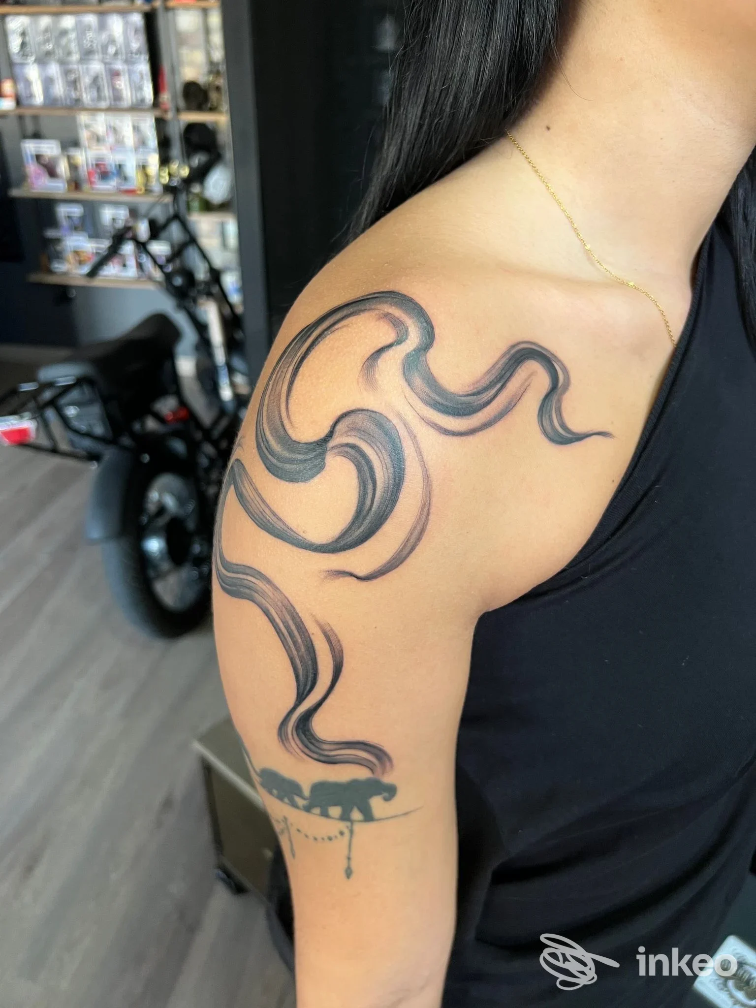 Tattoo sul braccio sinistro di una persona, disegno astratto in tonalità scure lungo la spalla, visibile esteso su bicipite; sfondo di studio Buena Suerte Tattoo e Piercing Studio a Pozzuoli.