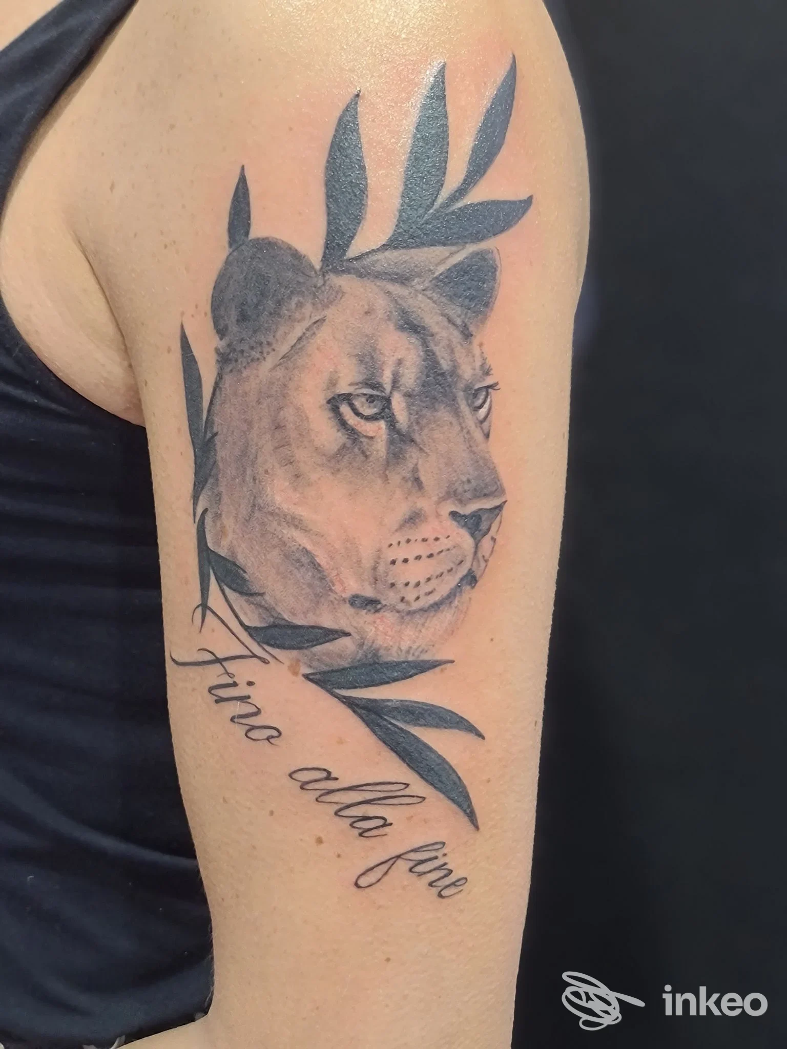 Tattoo sul braccio raffigurante una testa di leone in bianco e nero circondata da foglie, accompagnata da una scritta corsiva sul lato inferiore.