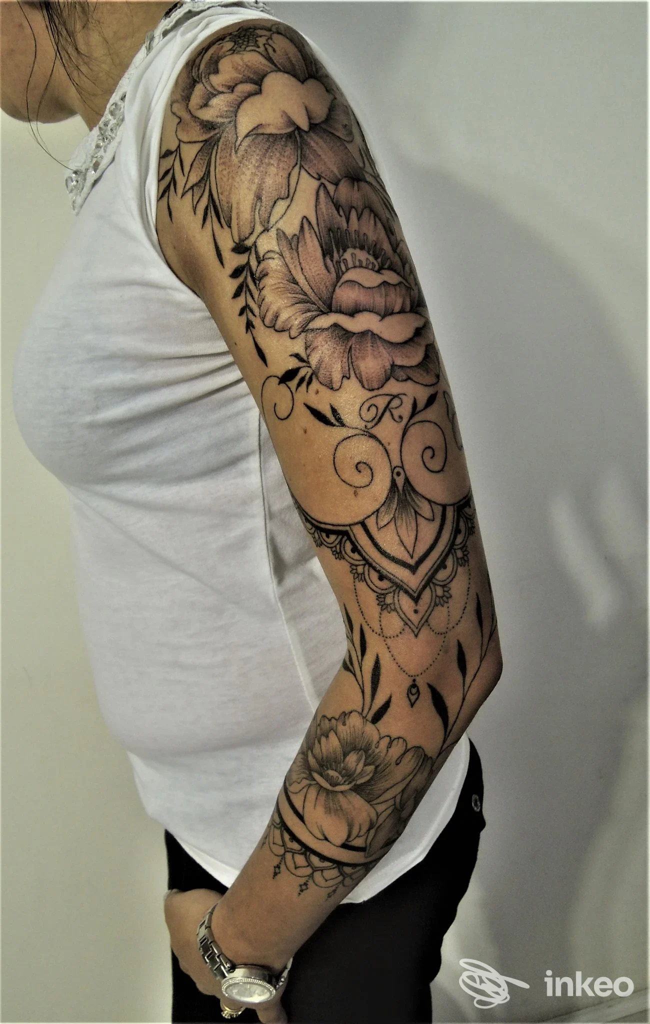 Braccio di una persona con grande tatuaggio floreale e motivi ornamentali, visibile sotto manica bianca, primo piano dal lato sinistro. Abito casual e sfondo neutro.