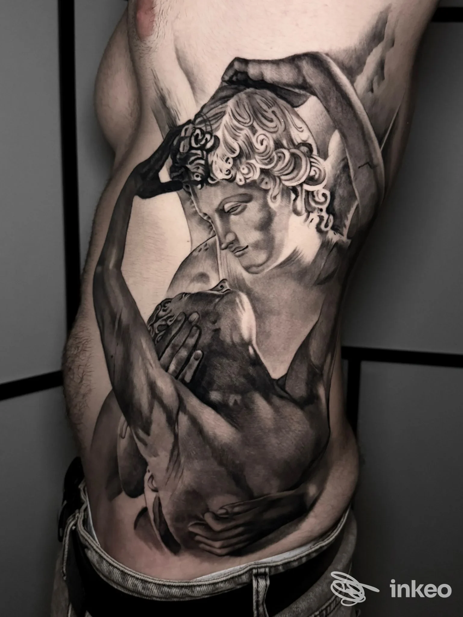 Tattoo realistico su torso raffigurante due figure abbracciate, stile scultoreo in bianco e nero.