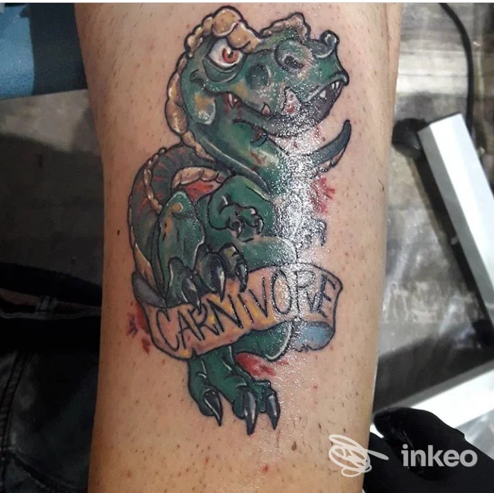 Tatuaggio realistico su pelle raffigurante un dinosauro verde con catena e banner che recita Carnivore; colori vividi e linee definite, nessun volto visibile.