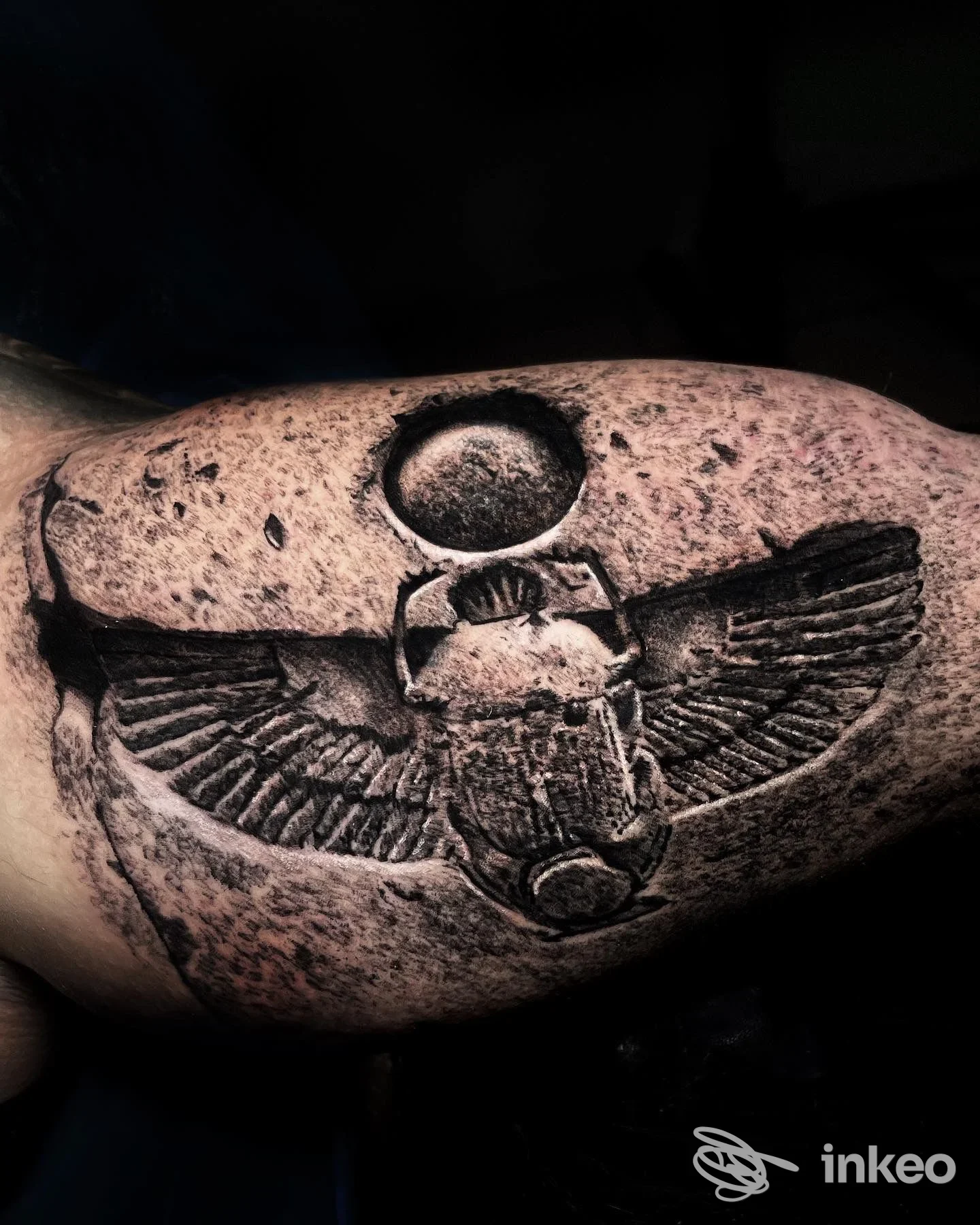Tattoo realistico di un amuleto o simbolo con ali su pelle, visibile su braccio/ferita, in tonalità scure, con sfondo scuro.