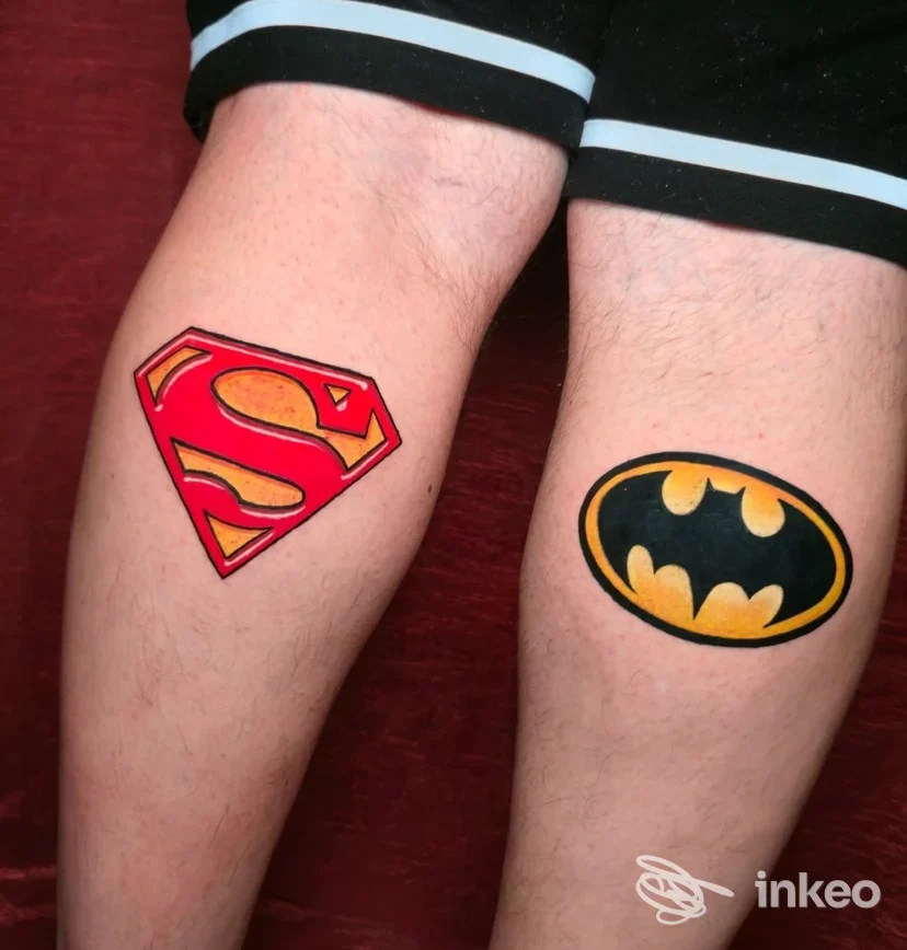 Due tatuaggi visibili sugli avambracci: sul braccio sinistro è raffigurato il logo di Superman con colori vivaci, sul braccio destro il logo di Batman su sfondo giallo e nero; nessun volto visibile, pelle chiara, sfondo rosso scuro.