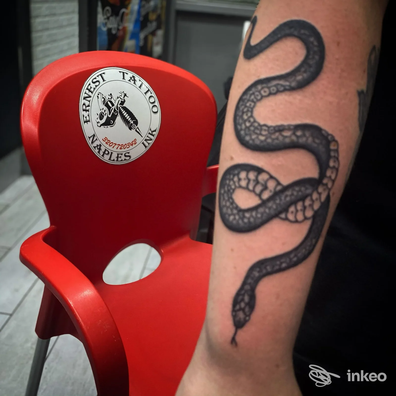 Braccio di una persona con tatuaggio serpente scuro ben definito, vicino a una sedia rossa in un salone.