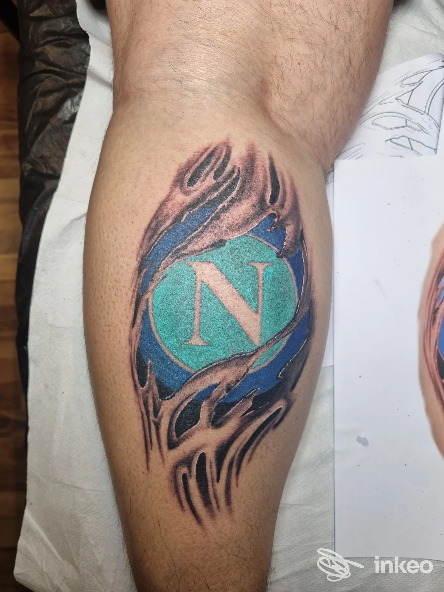 Tattoo colorata su avambraccio con sfondo turchese e una lettera N al centro, circondata da elementi grafici dorati/neri.