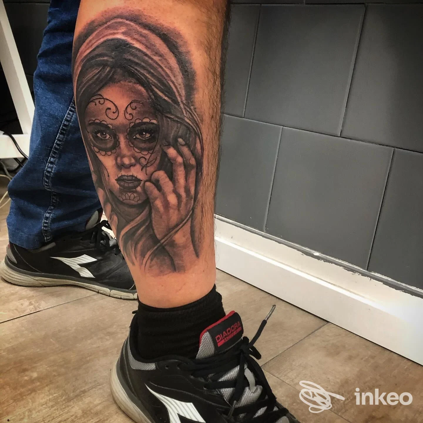 Tattoo su gamba visibile: ritratto in stile sugar skull di una donna, con dettagli scuri e linee nette; nessun volto intero mostrato, superficie cutanea ben illuminata, indossa una sneakers nera.