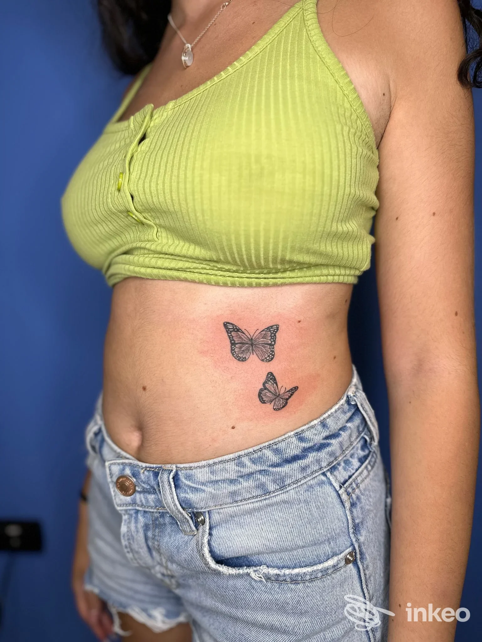 Due tatuaggi a farfalla sulla parte laterale del ventre di una persona, con top verde e jeans chiari, non visibile volto completo.