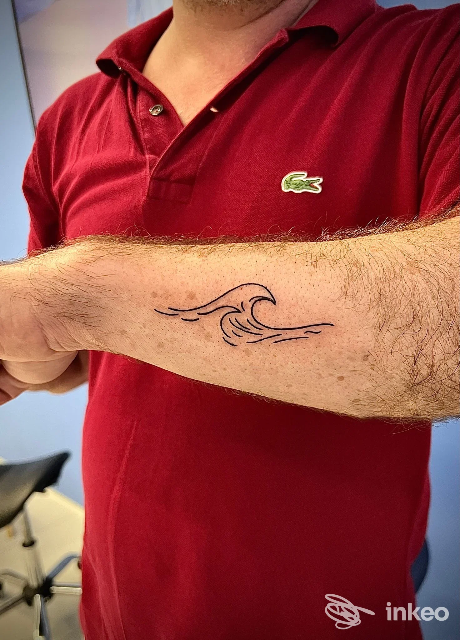 Braccio di uomo con tatuaggio ad onda stilizzata in nero sulla pelle, visibile sotto manica polo rossa, sfondo interno studio.