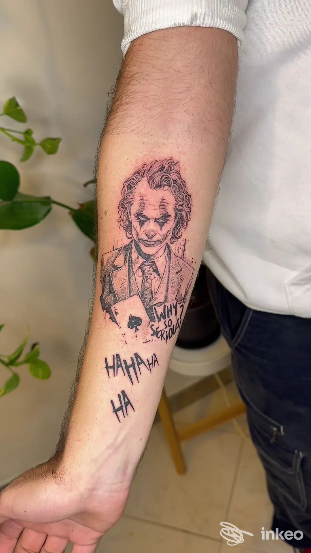 Braccio sinistro mostrando un tatuaggio con il Joker in stile tratto a china, con testo ha ha ha e Why so serious, su pelle chiara. stile dark line work.
