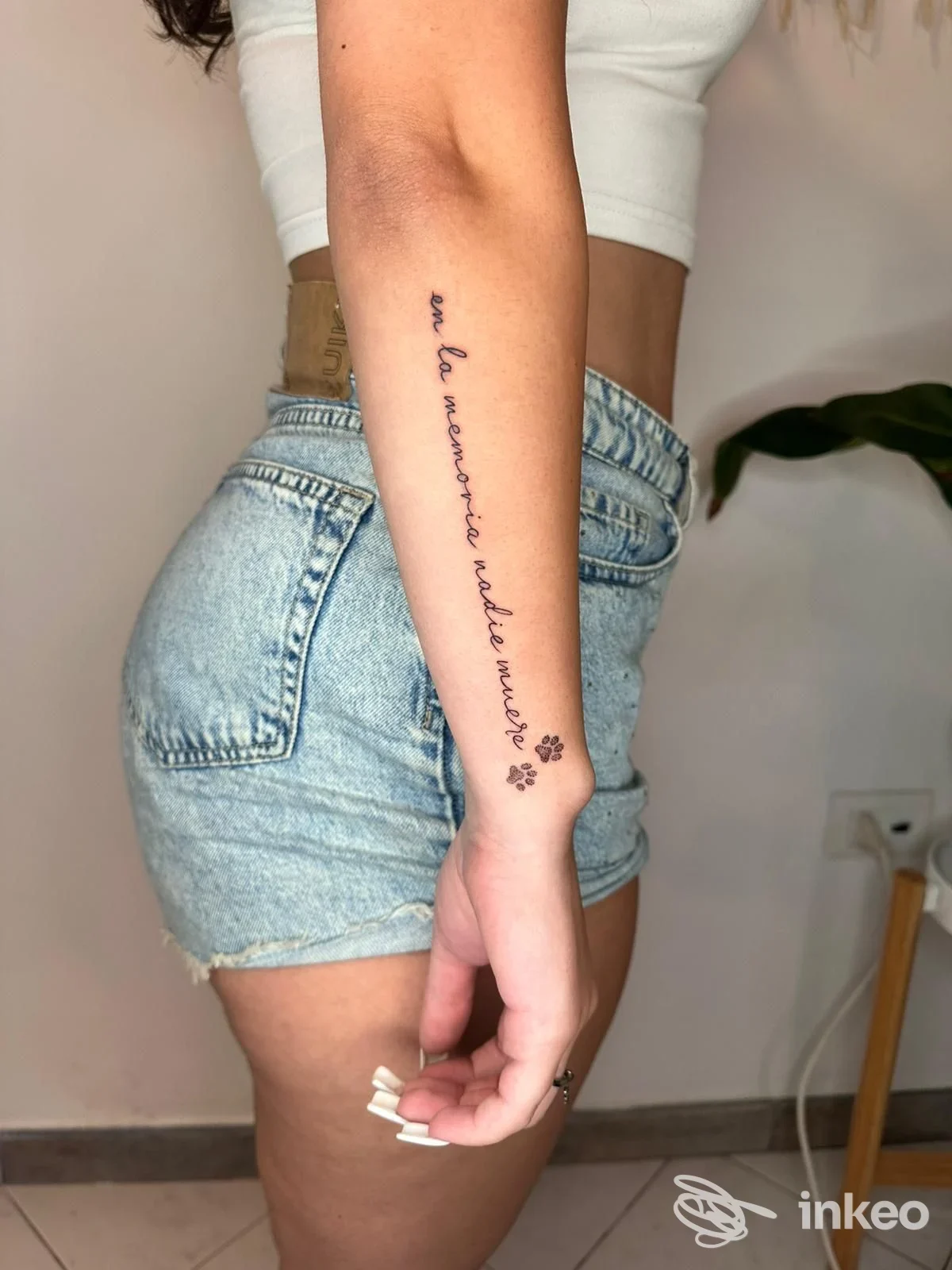 Avambraccio sinistro visibile con tatuaggio testo in corsivo lungo la pelle, accompagnato da piccole impronte, indossando una maglietta corta e shorts di jeans in un ambiente domestico.