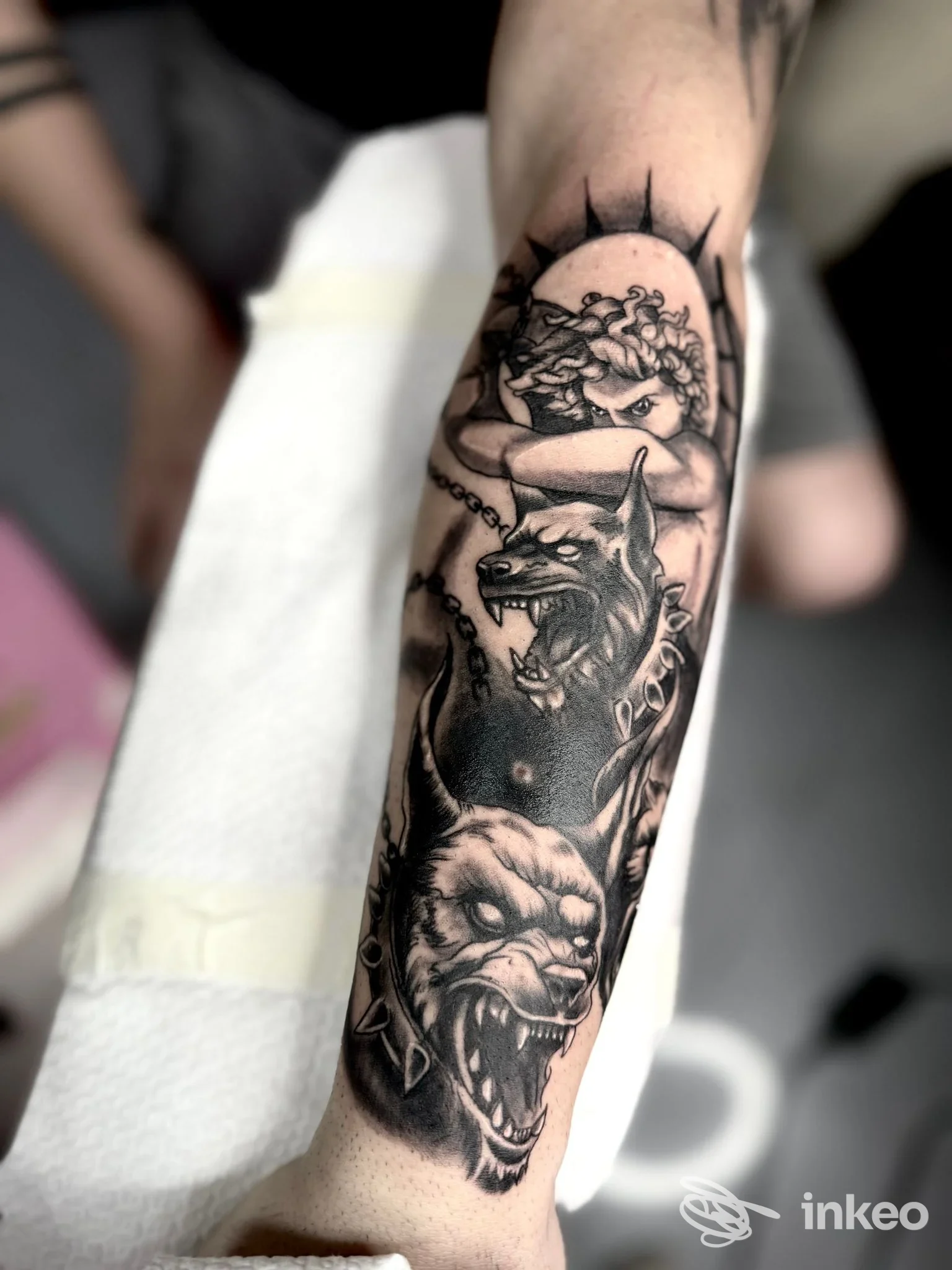 Tatuaggio in bianco e nero su avambraccio: figura di cane lupo con zanne e catena, accompagnato da una figure laterale, tutto in stile realistico.