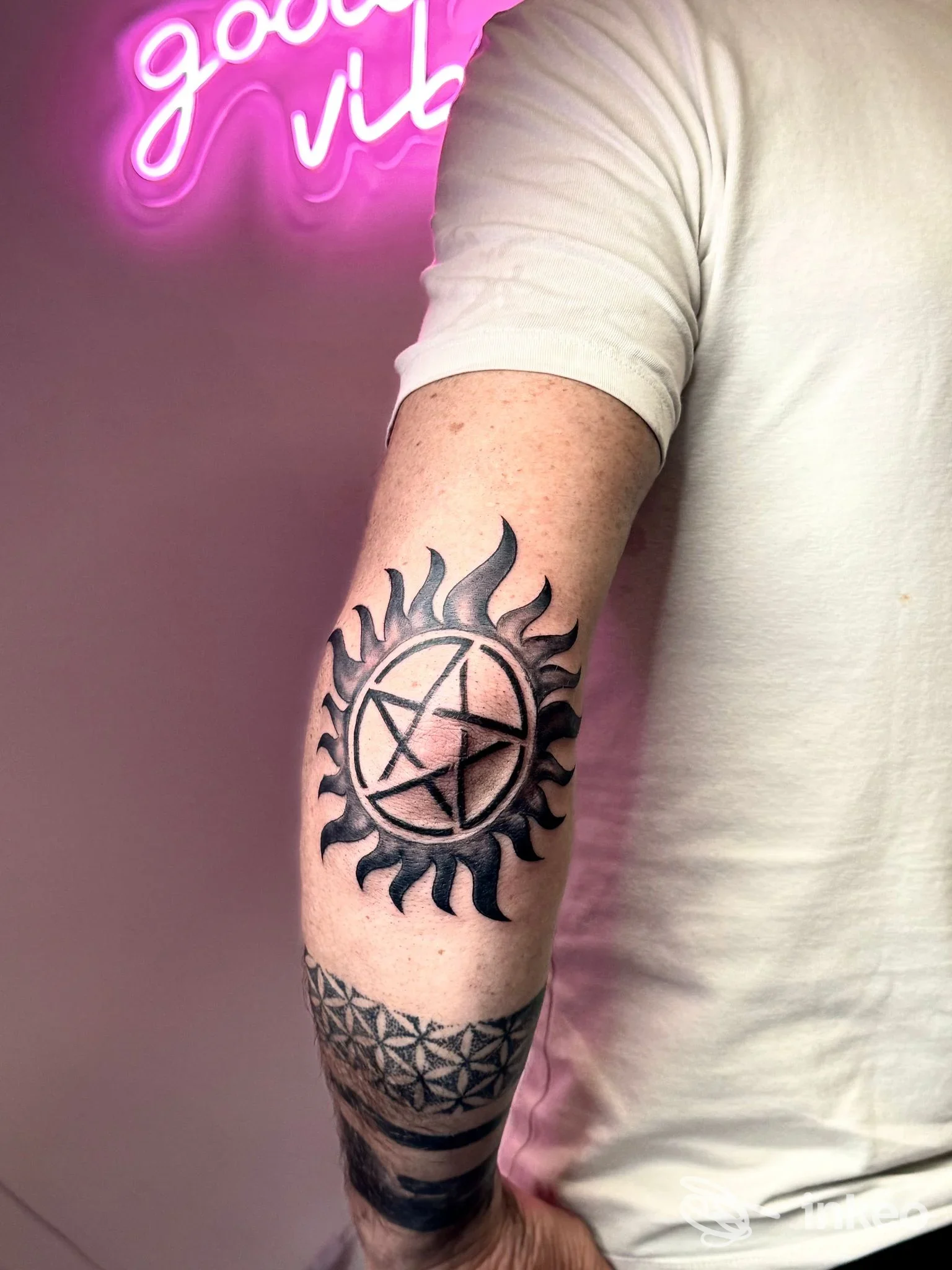 Braccio di una persona con tatuaggio nero a tema circolare con simboli simmetrici, illuminato da luci rosa, sfondo neutro; studio Alex Abate tattoo ink, Aversa.