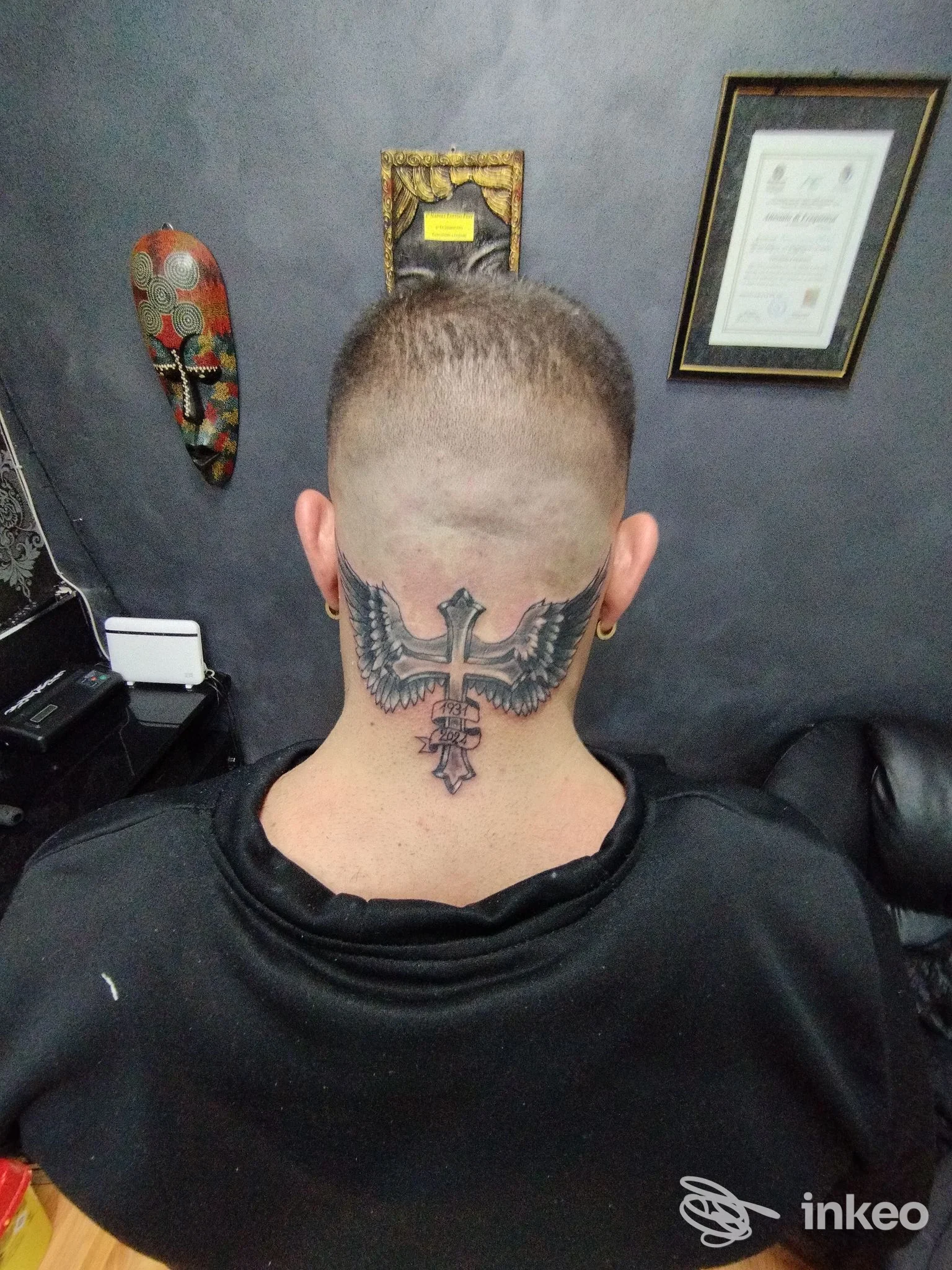 Tattoo sul retro del collo: croce con ali stilizzate, scuro su pelle chiara, visibile da dietro la nuca, di fronte a pareti e cornici in studio.