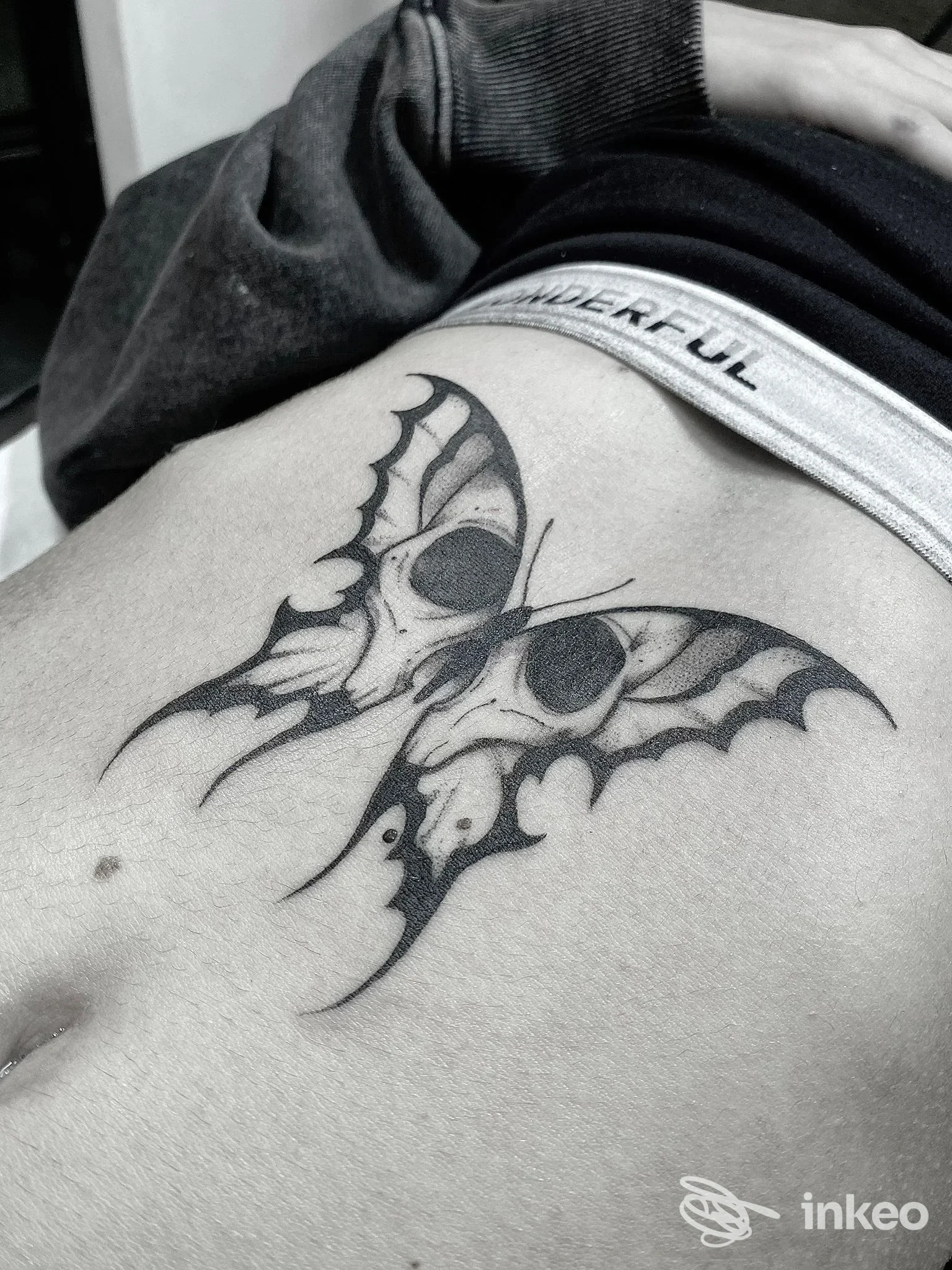 Tattoo reale su pelle: braccio o fianco con disegno di pipistrello in bianco e nero, tonalità scure, bordo elastico visibile. Estremità della ferita non visibile.